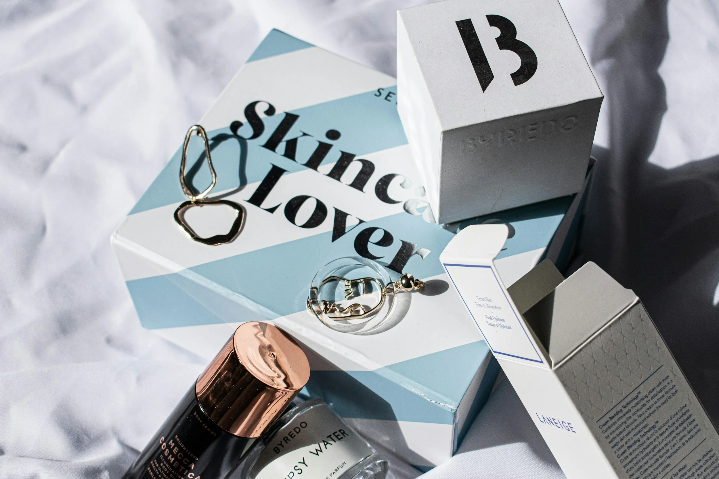 A flat lay of skincare and jewelry items on a white wrinkled surface. Contains a blue and white box with the words 'Skincare Lover', a skincare bottle with a rose gold cap, a small perfume bottle, and jewelry pieces including a ring and a tear-shaped pendant.