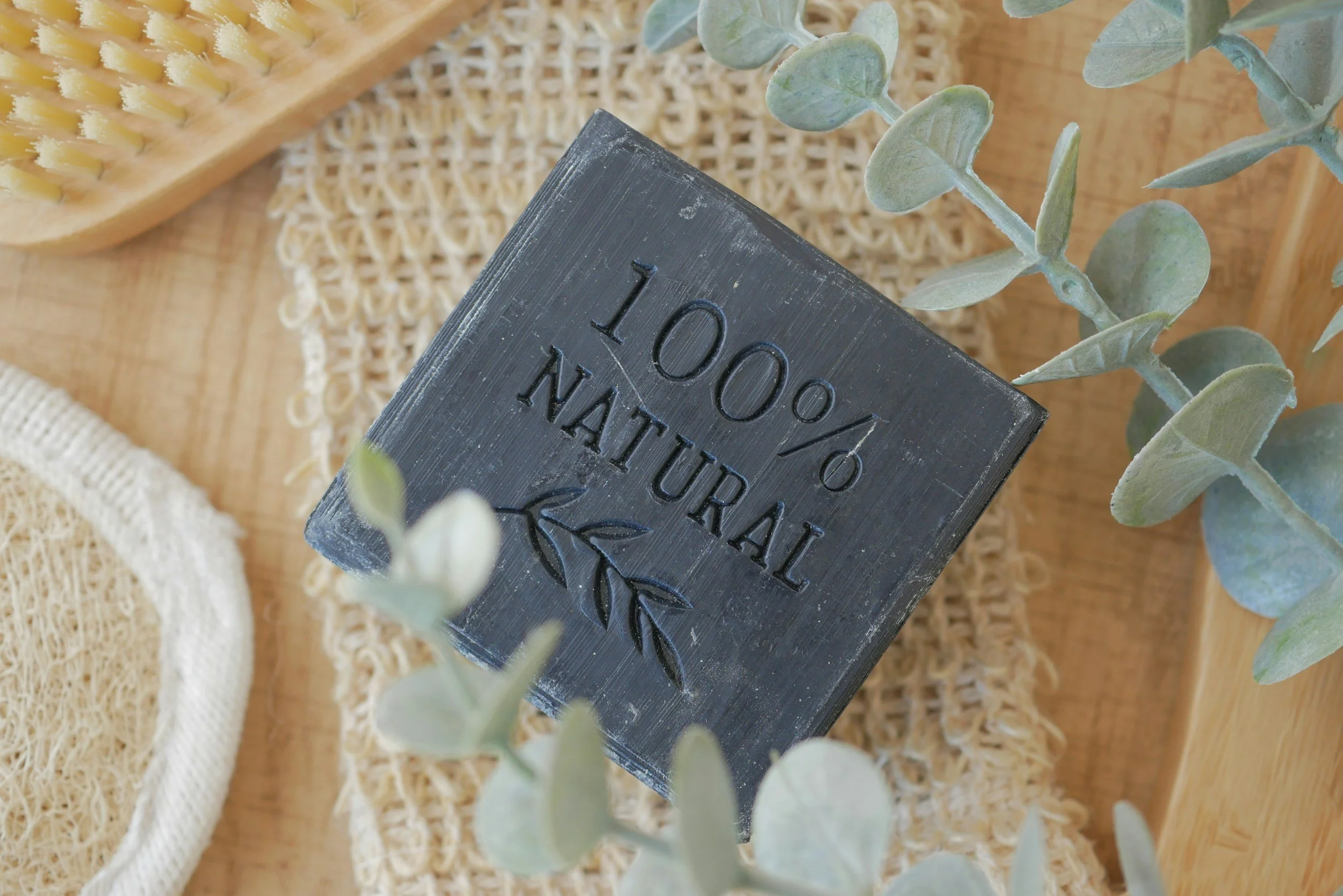 A black wooden sign with the text '100% NATURAL' engraved on it, surrounded by eucalyptus leaves and natural woven and wooden items.