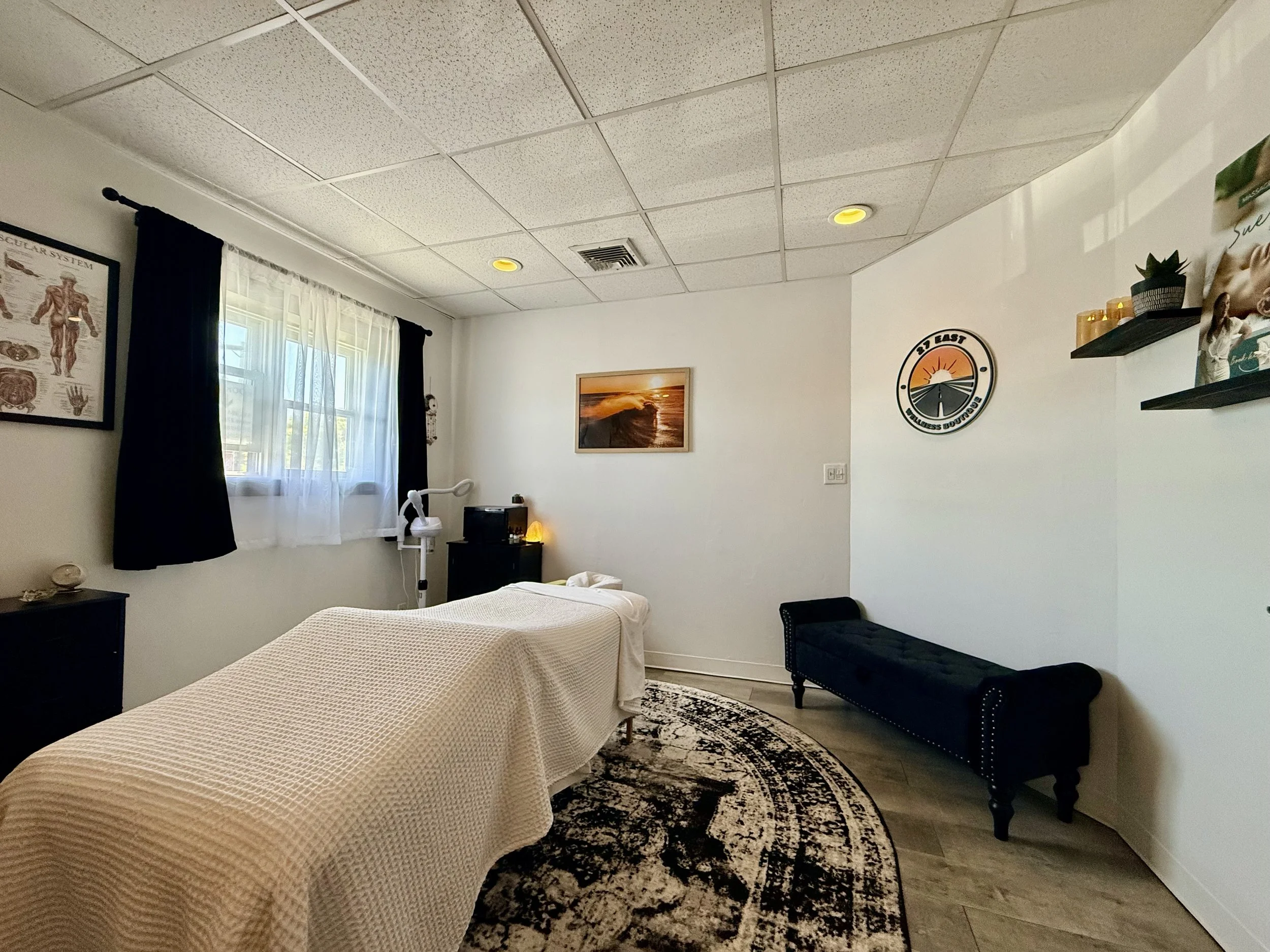 A cozy bedroom with two beds covered in beige bedding, a black and white patterned rug, wall art including a sunset photo and a sport logo, shelves with plants, candles, and a photograph, a bedside table with a lamp, and a window with white sheer curtains and black drapes.