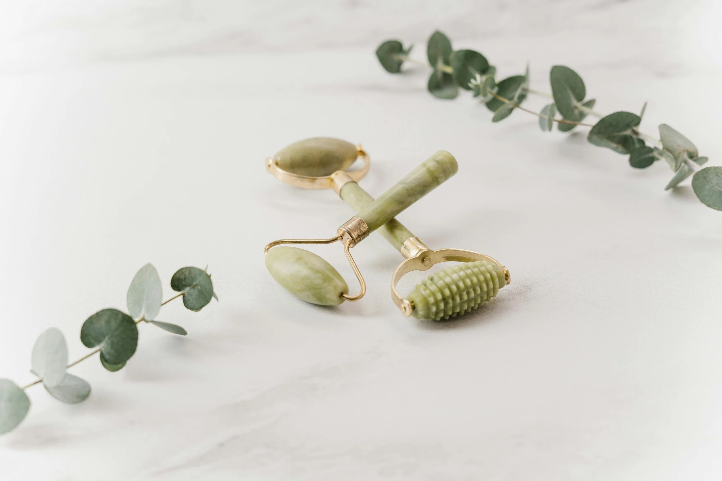 A green jade facial roller, a green jade gua sha tool, and a green silicone facial scrubber are arranged on a white surface, with a eucalyptus branch nearby.