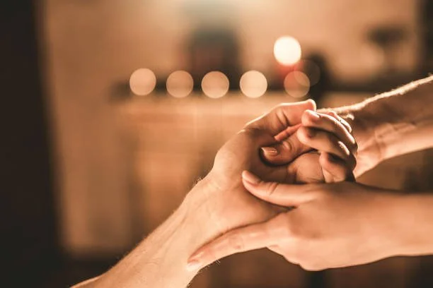 Two people holding hands, one person clasping the other's hand gently in a warm, cozy setting with blurred background lights.