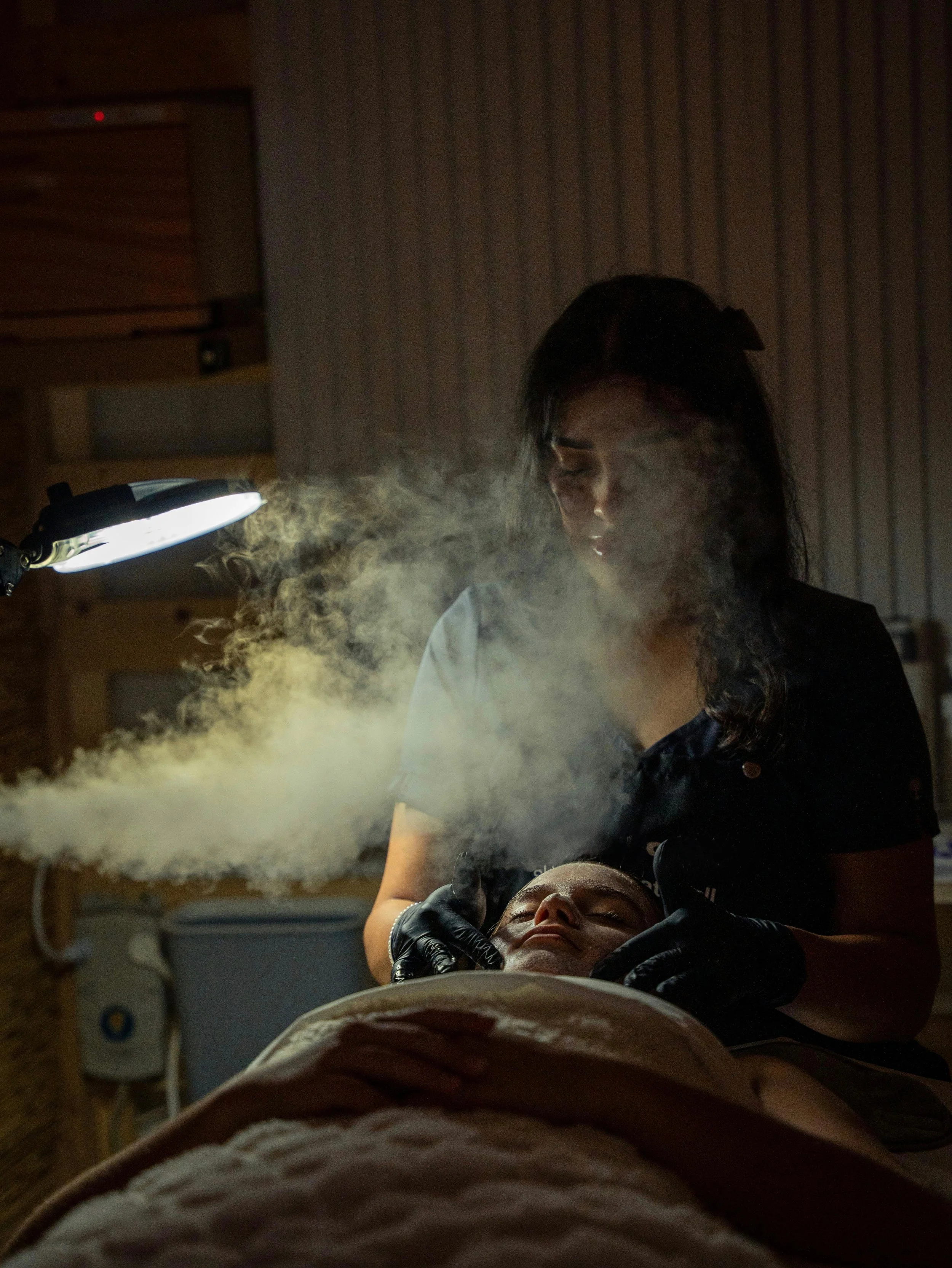A woman is performing a cosmetic procedure on a person's face in a dimly lit room, with steam or vapor rising around them, illuminated by a bright lamp.