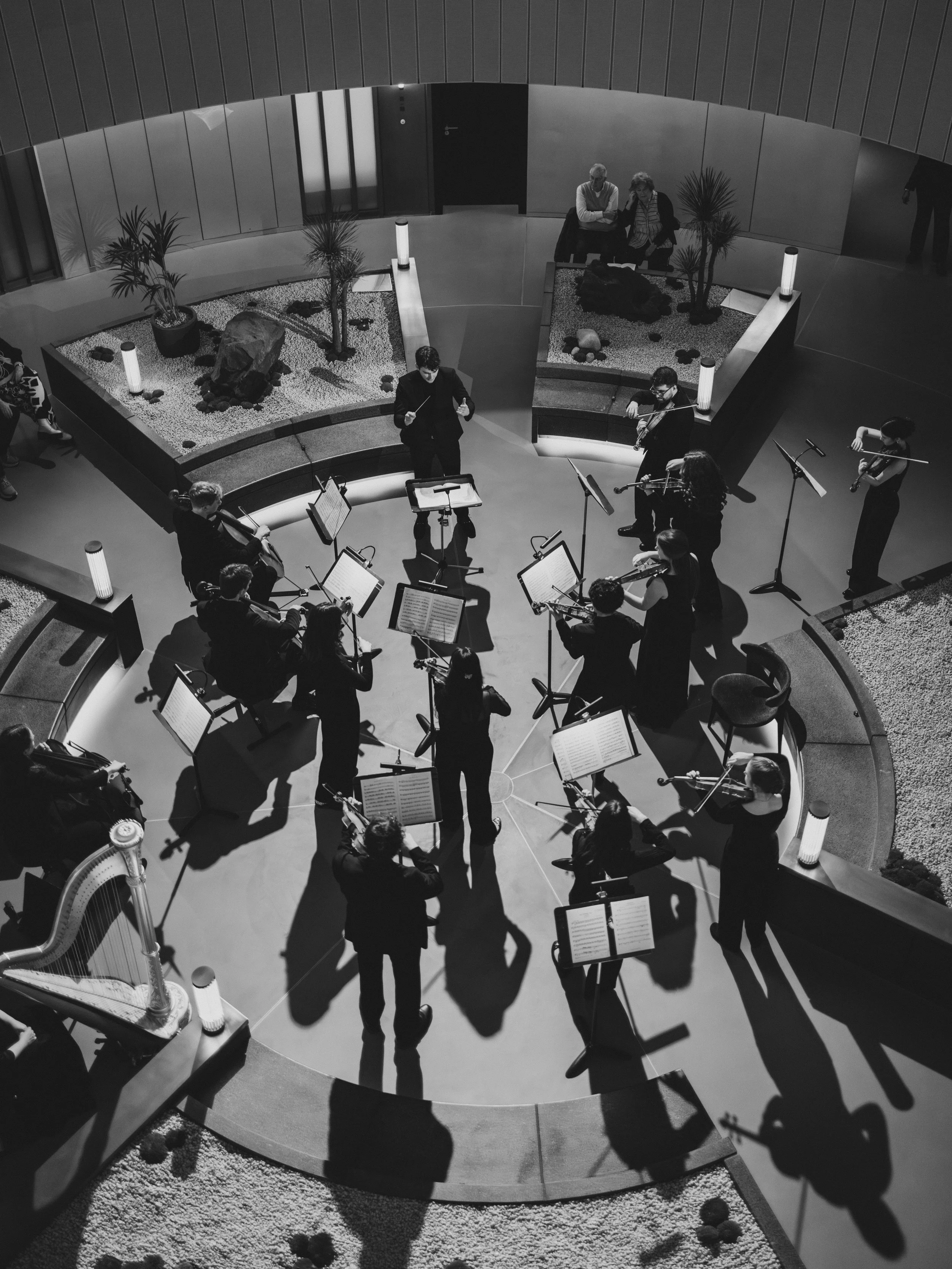 A group of orchestral musicians performing in a semicircular arrangement in an indoor atrium with decorative plants, a harp, and two people sitting on a bench observing.