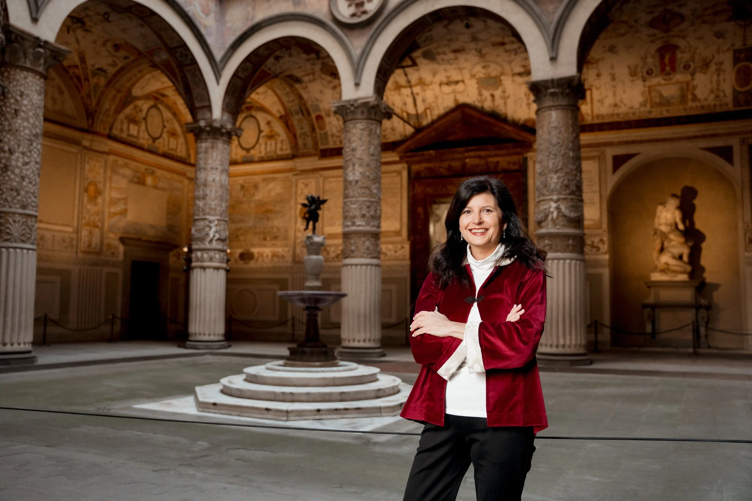 A woman in a red jacket standing in an ornate historic building with columns and sculptures, smiling with arms crossed.