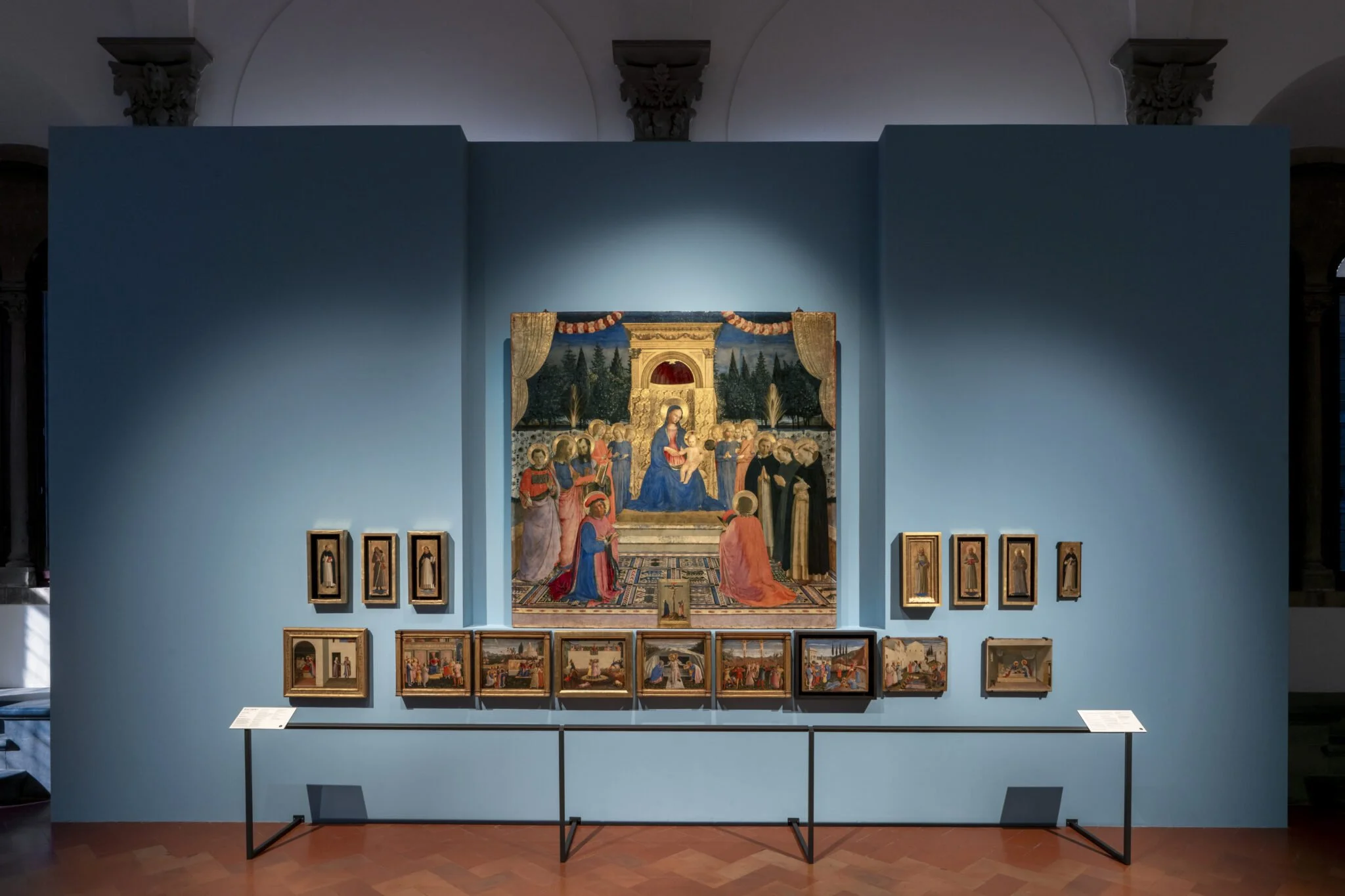 Art exhibit featuring a large religious painting of the Madonna and Child surrounded by smaller framed artworks on a bright blue wall in a museum.