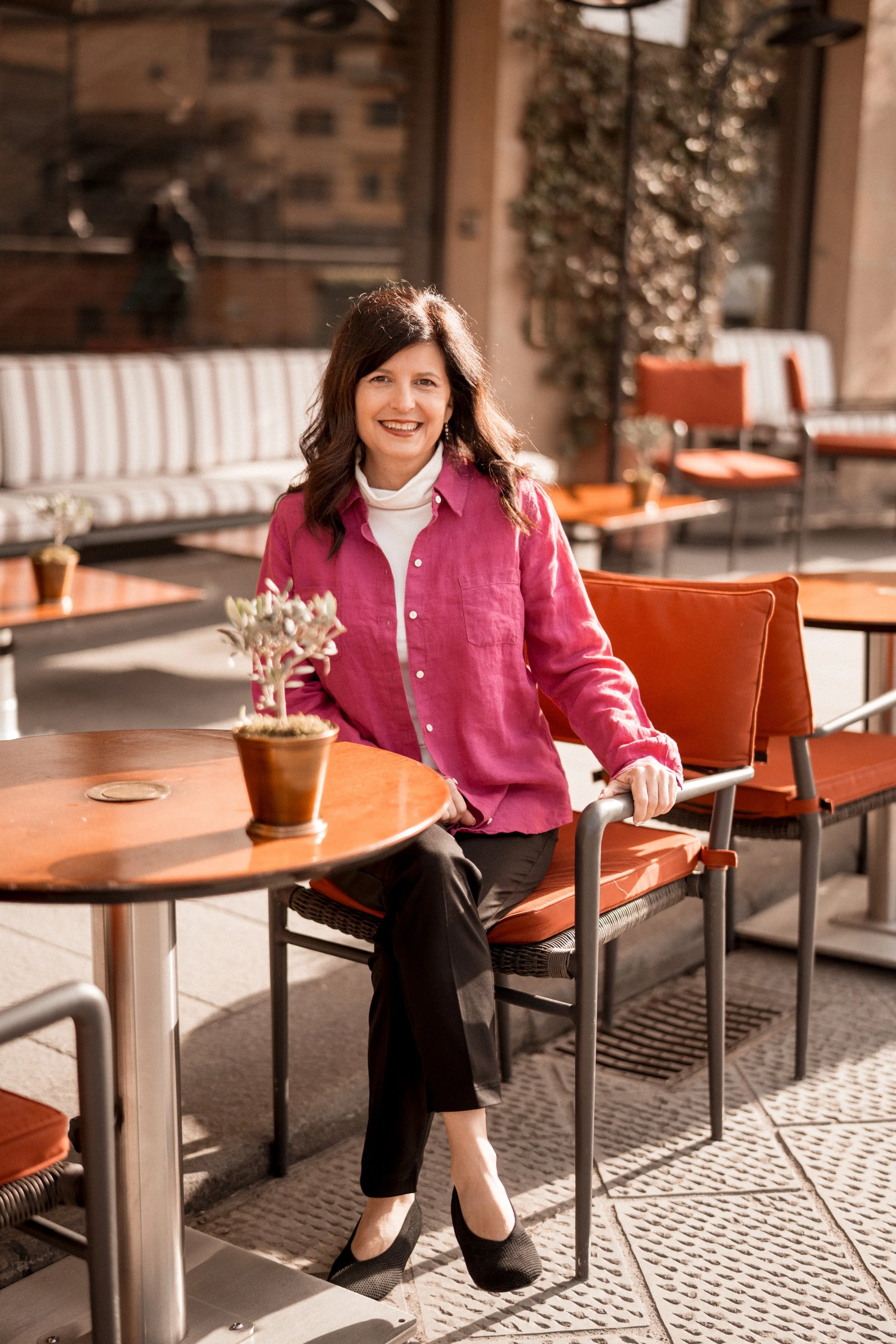 A woman with dark, shoulder-length hair smiles while sitting at an outdoor patio table. She is wearing a white turtleneck under a pink button-up shirt and black pants. There is a small potted plant on the table and empty chairs and seating in the background.