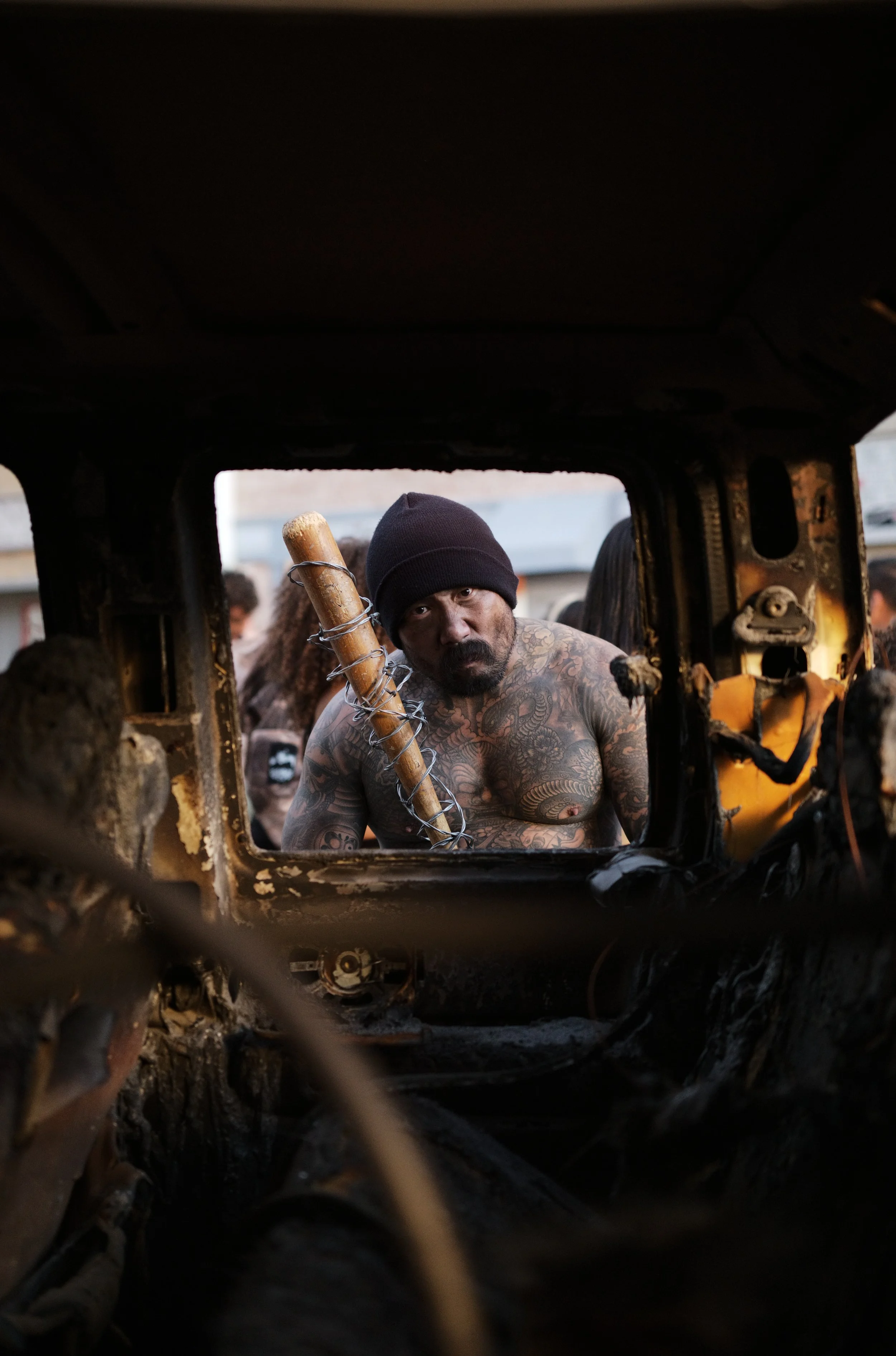 A shirtless man with tattoos and a beanie holding a machete, seen through a damaged and burnt vehicle window.