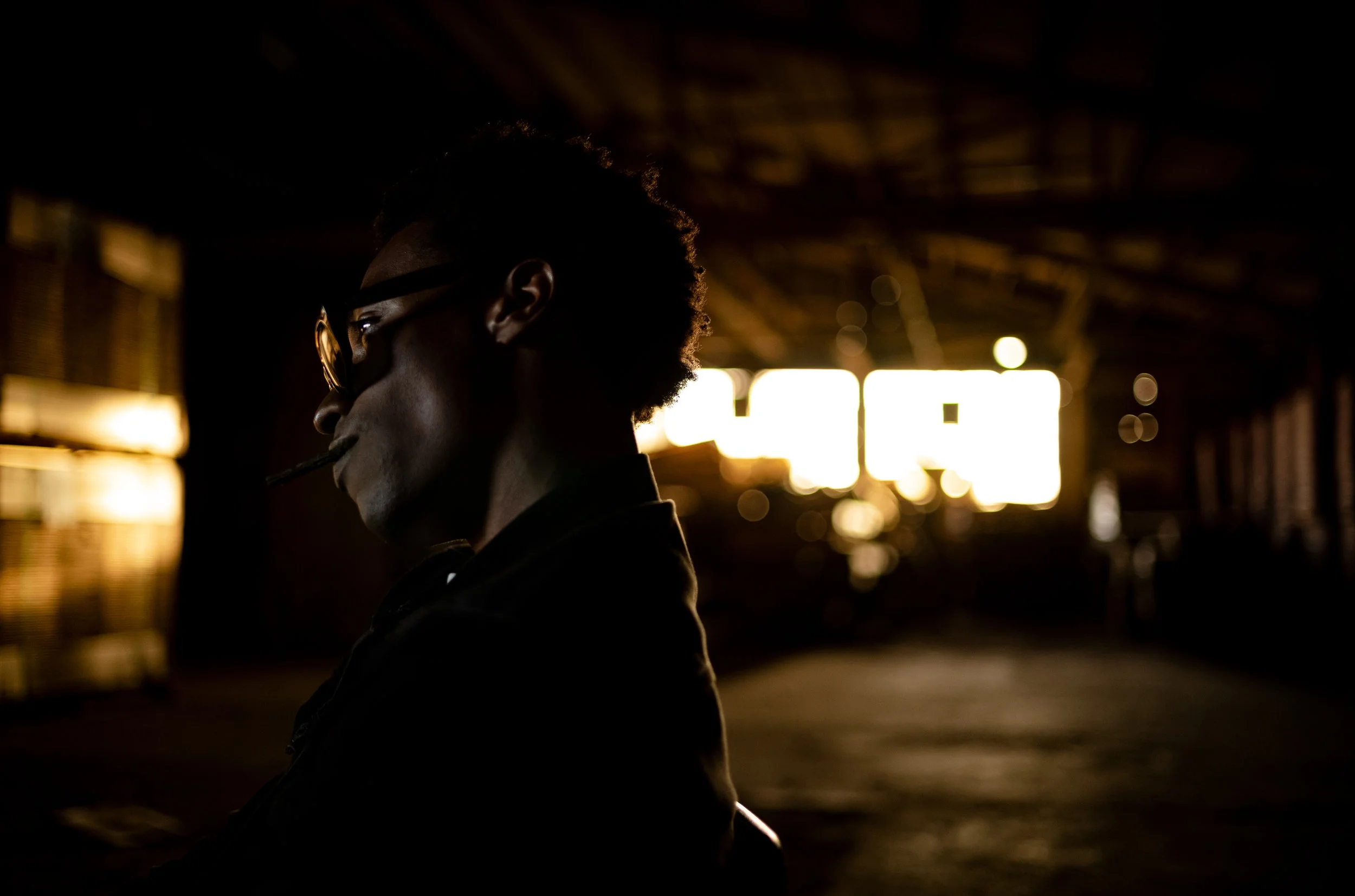 Silhouette of a man with glasses smoking a cigar in an industrial or warehouse setting with bright sunlight or artificial light in the background.