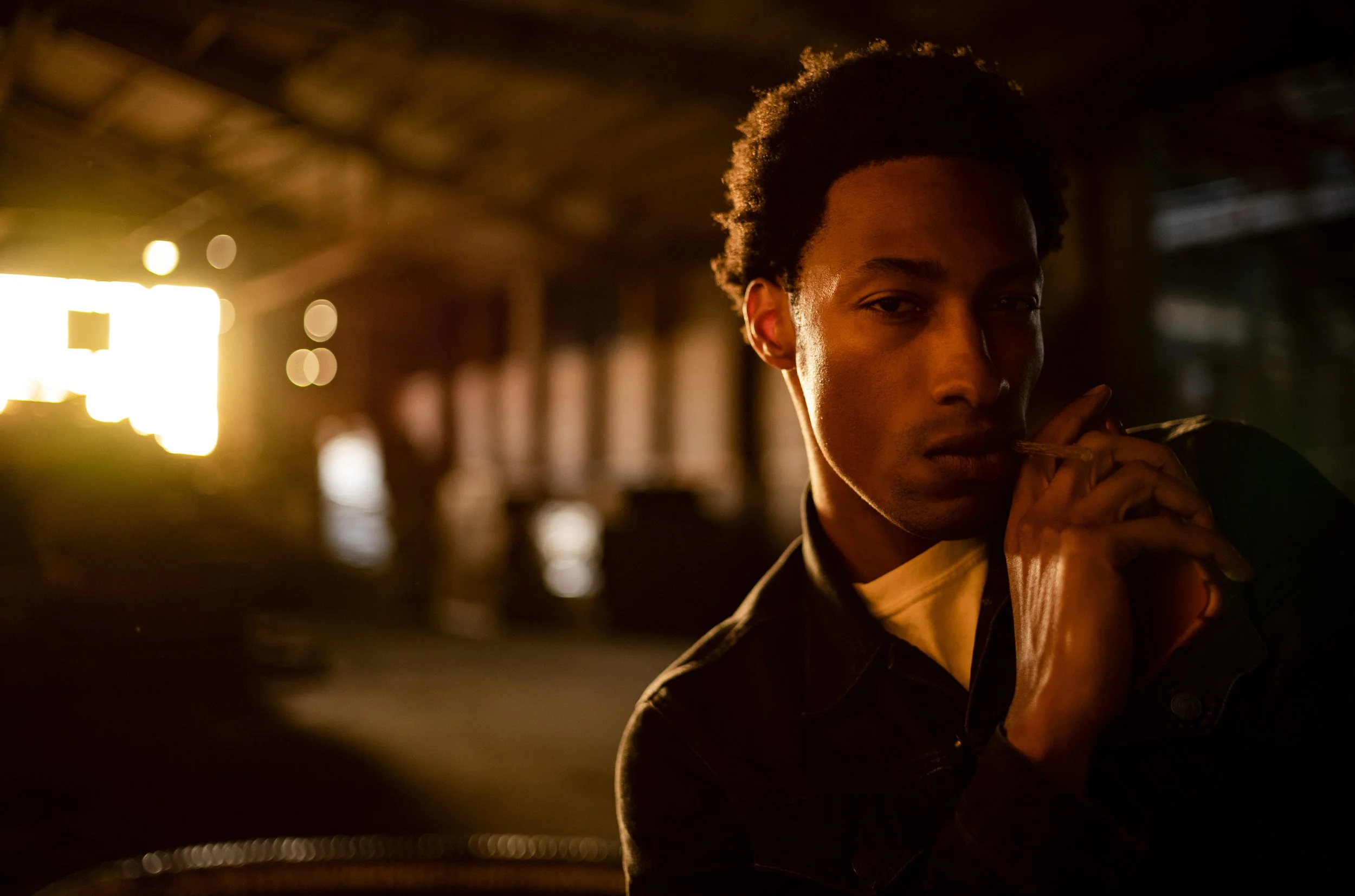 A young man with dark, curly hair pose in a dimly lit, industrial setting, holding a cigarette near his face, with warm backlighting creating a silhouette effect and blurred background.