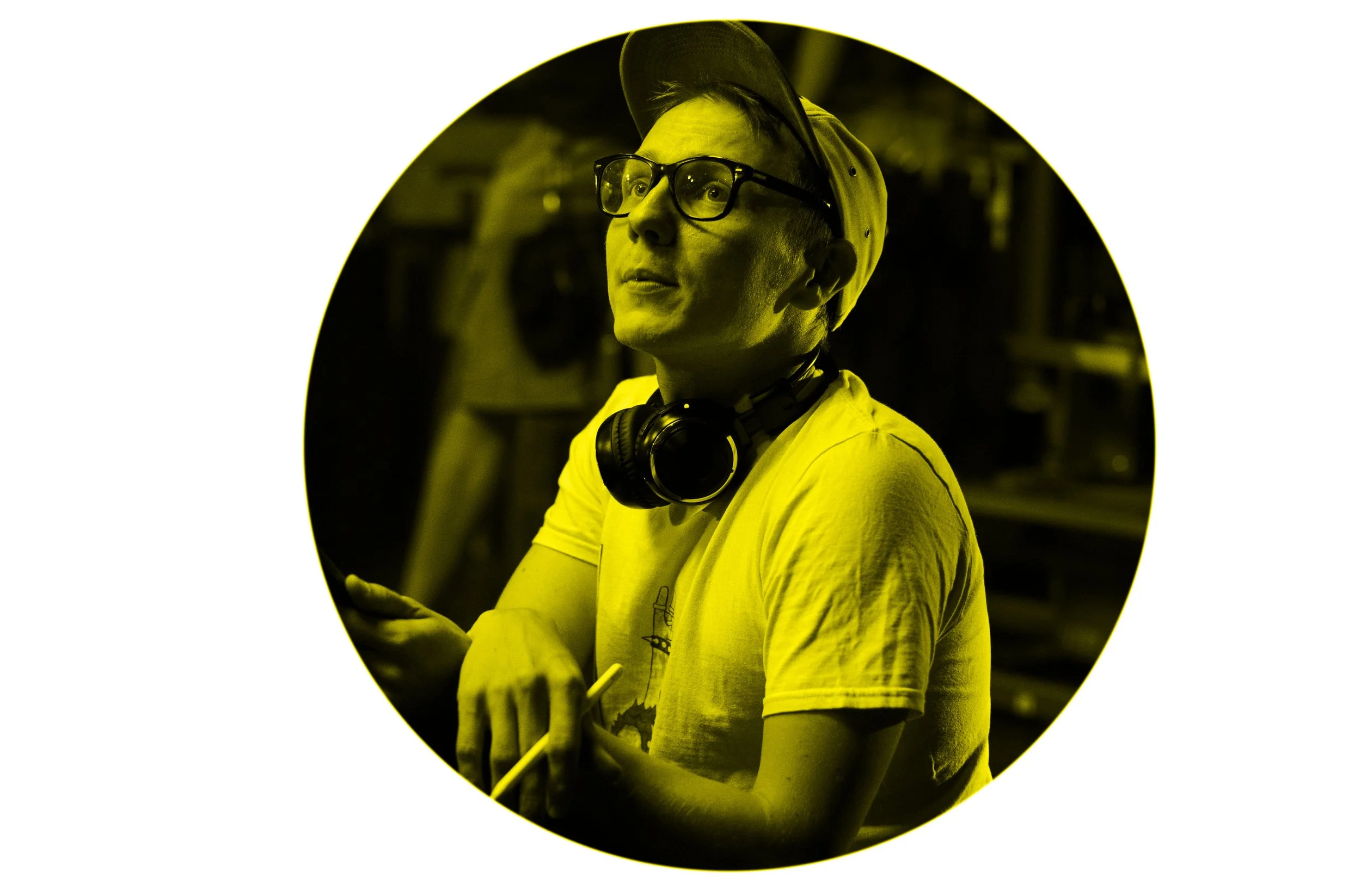 A young man wearing glasses, a cap, and headphones around his neck, holding a stylus pen, looks upward with a thoughtful expression, in front of shelving and machinery.