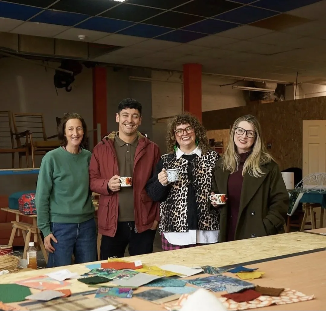 Four people standing in a room, smiling and holding mugs, with a table of fabric swatches and crafting supplies in the foreground.