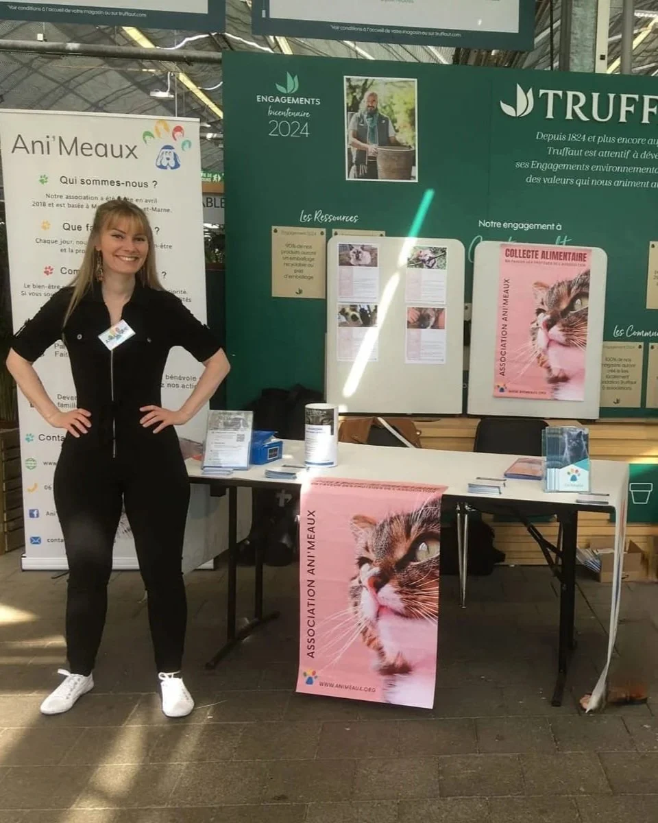Une femme souriante pose à côté d'un stand d'une association animale, avec des affiches et des brochures sur la protection des animaux, notamment un panneau avec un gros chat et le logo de l'association Ani'Meaux.