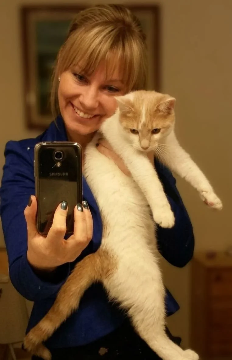 Une femme souriante tenant un chat roux et blanc devant un miroir pendant qu'elle se prend en selfie avec un téléphone Samsung.