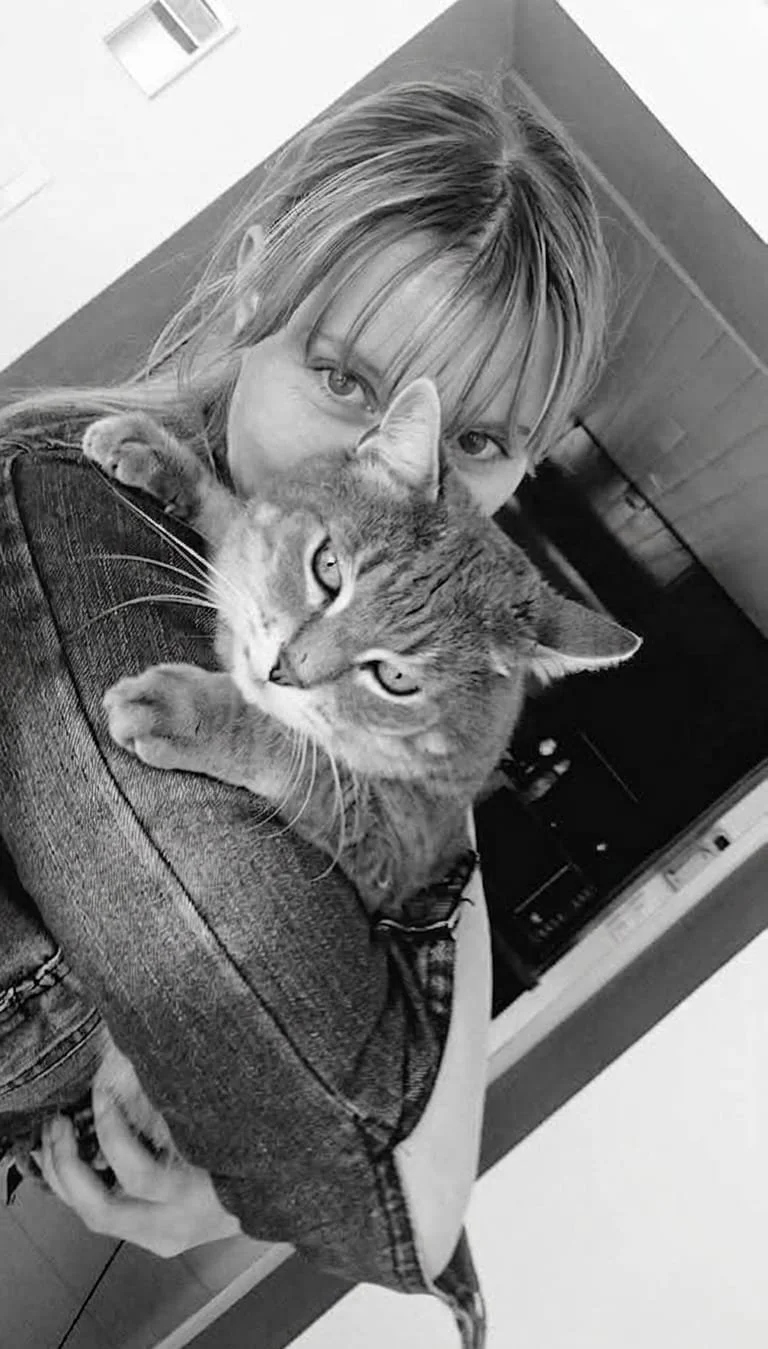 Une femme avec des cheveux attachés, portant une veste en jean, tient un chat tigré dans ses bras, en l'embrassant, les deux regardent l'appareil photo. La photo est en noir et blanc.