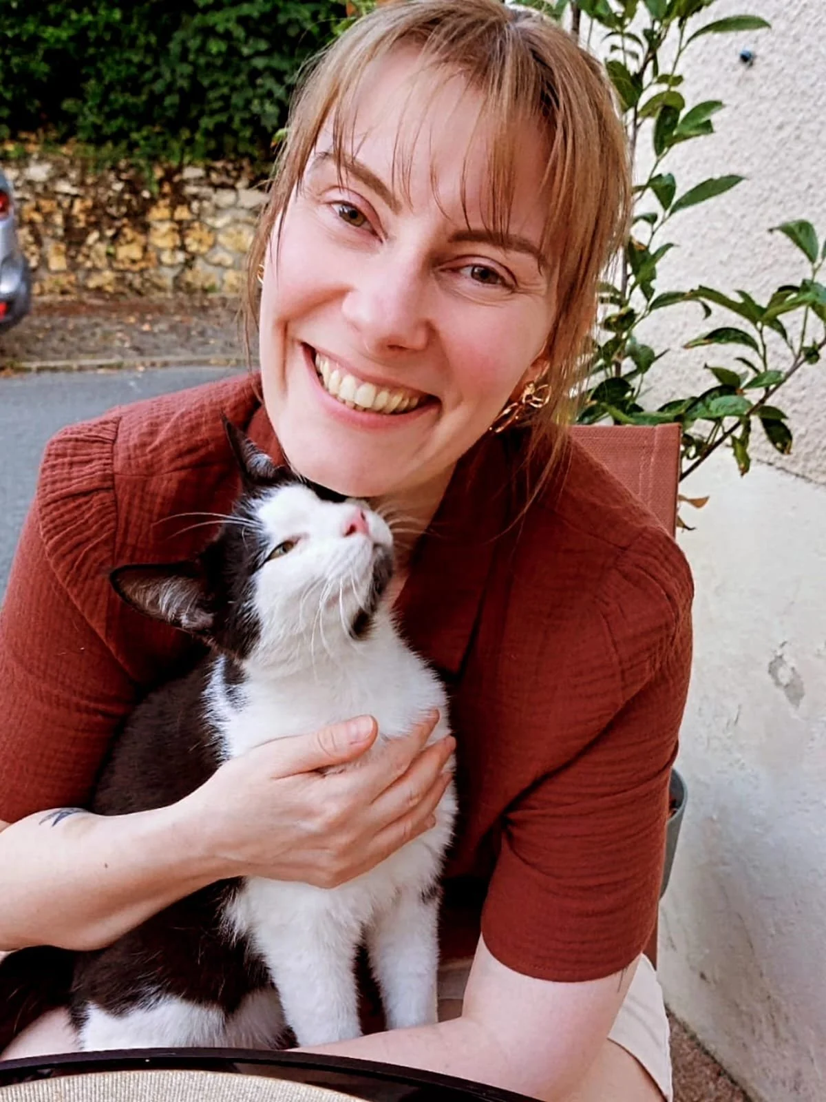 Une femme souriante tient un chat noir et blanc près de son visage en extérieur.