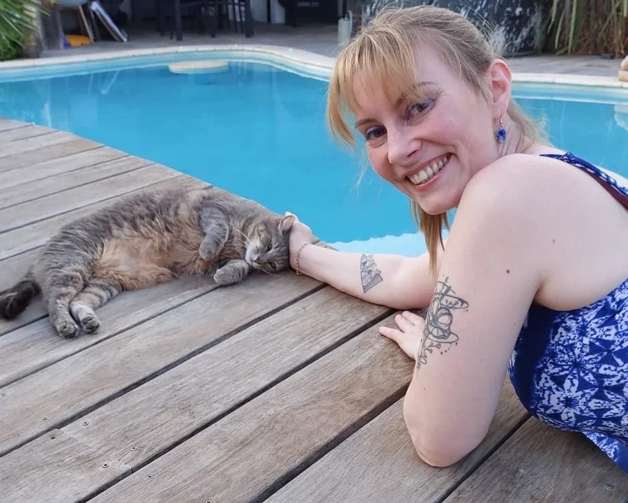 Une femme souriante qui fait un câlin à un chat gris allongé sur une terrasse en bois près d'une piscine.