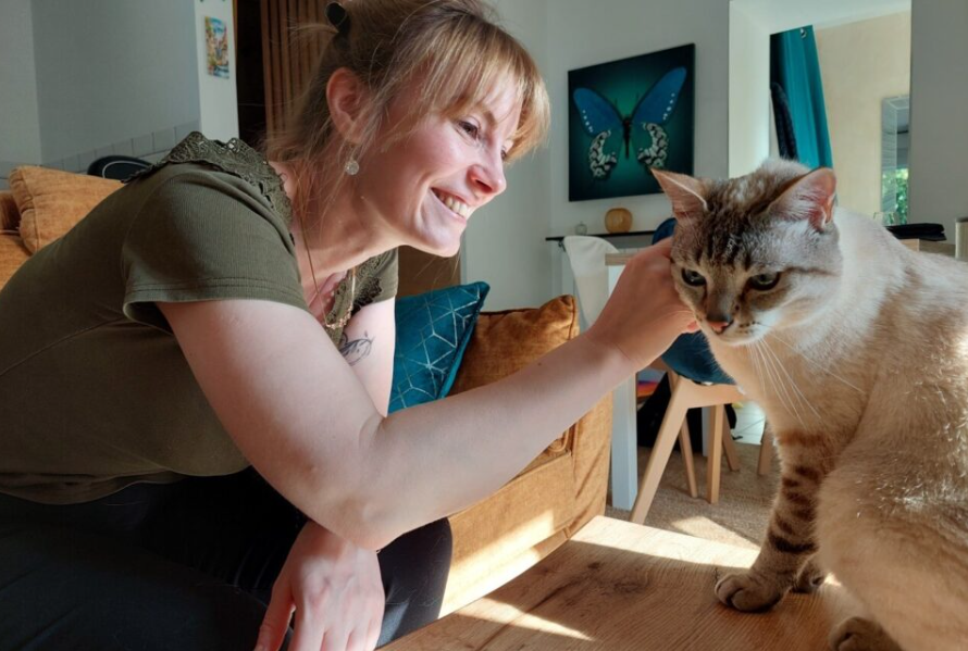 Une femme souriante caresse un chat domestique dans un salon bien éclairé, avec un tableau de papillons en arrière-plan.