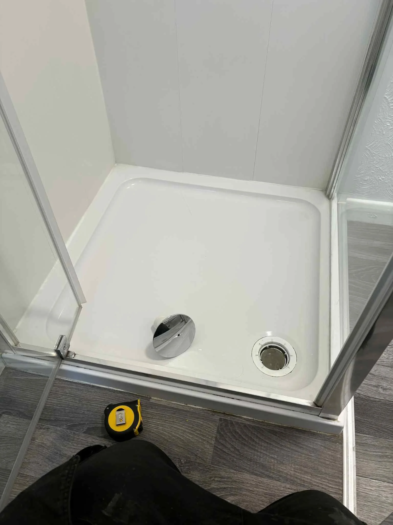 Empty shower stall with a drain, a reflective disc, a tape measure on dark wood floor, and a person's knee in foreground.