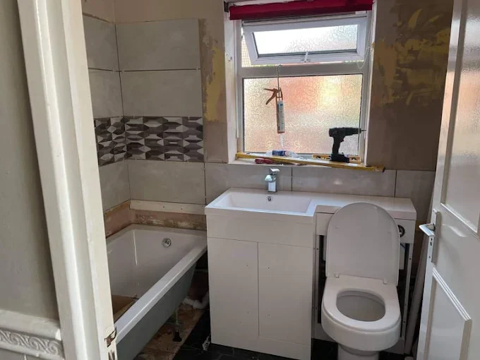 A small bathroom with unfinished tiling on the walls, a bathtub, a small sink cabinet, and a toilet, with a window above the sink and various construction tools on the windowsill.