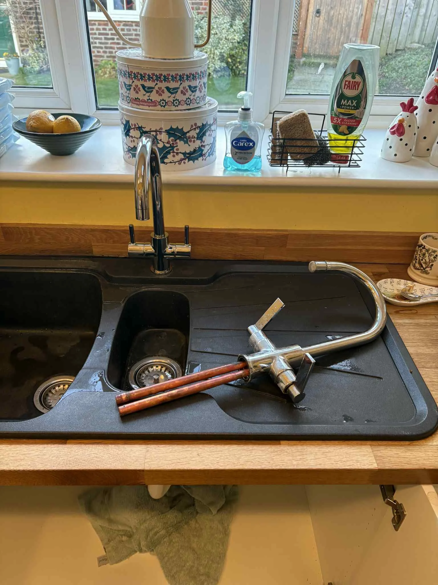 Kitchen sink with removed plumbing parts lying on the black sink surface, with a faucet above the sink, and a window above the sink showing a garden outside. The window sill holds a bowl of lemons, decorative tins, a soap dispenser, a sponge, and chicken-shaped ceramic containers.