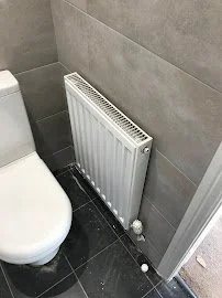 A white radiator heater mounted on a gray tiled bathroom wall next to a toilet with a black tiled floor.