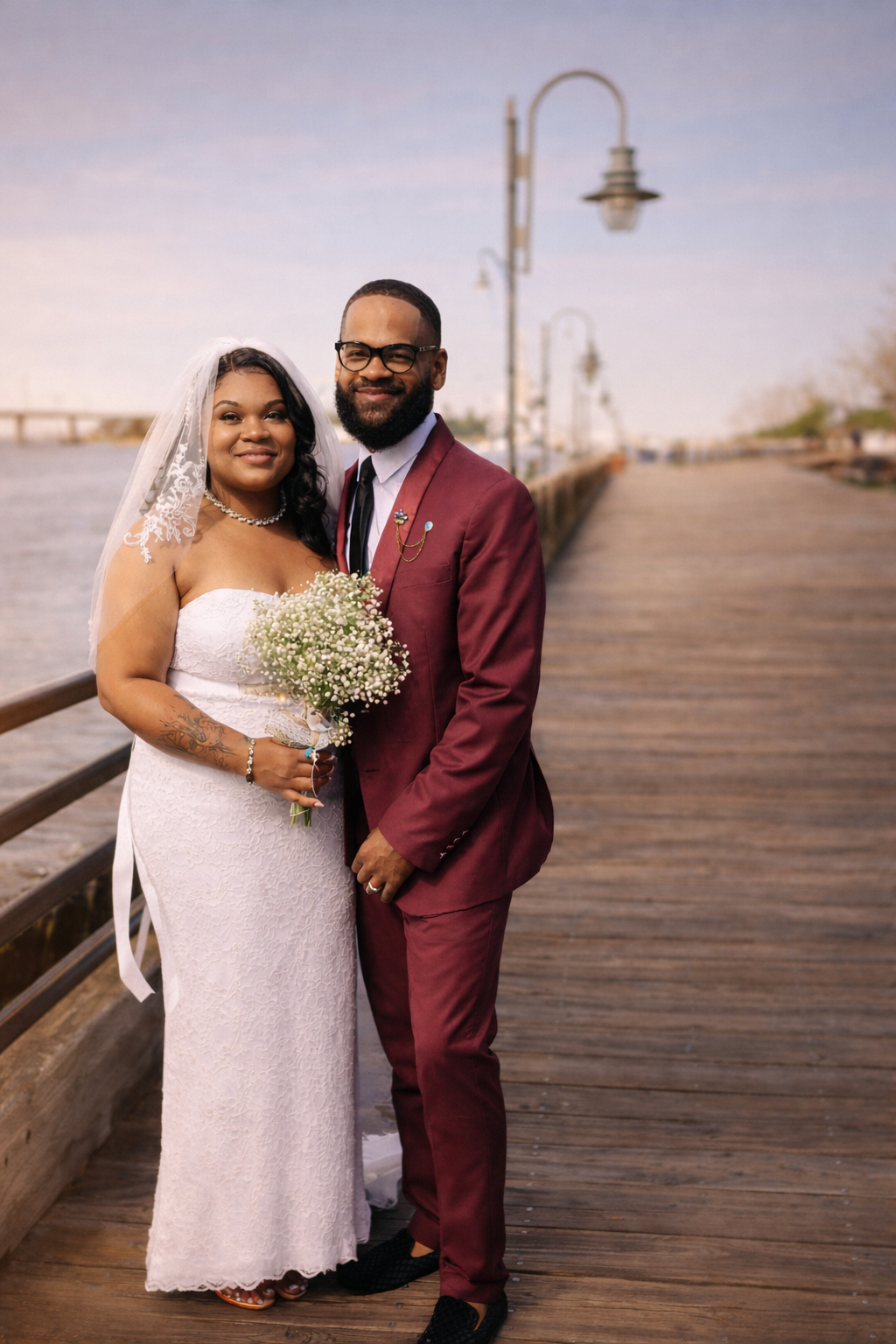 A newlywed couple standing on a wooden pier beside a body of water during sunset, dressed in wedding attire, smiling at the camera.