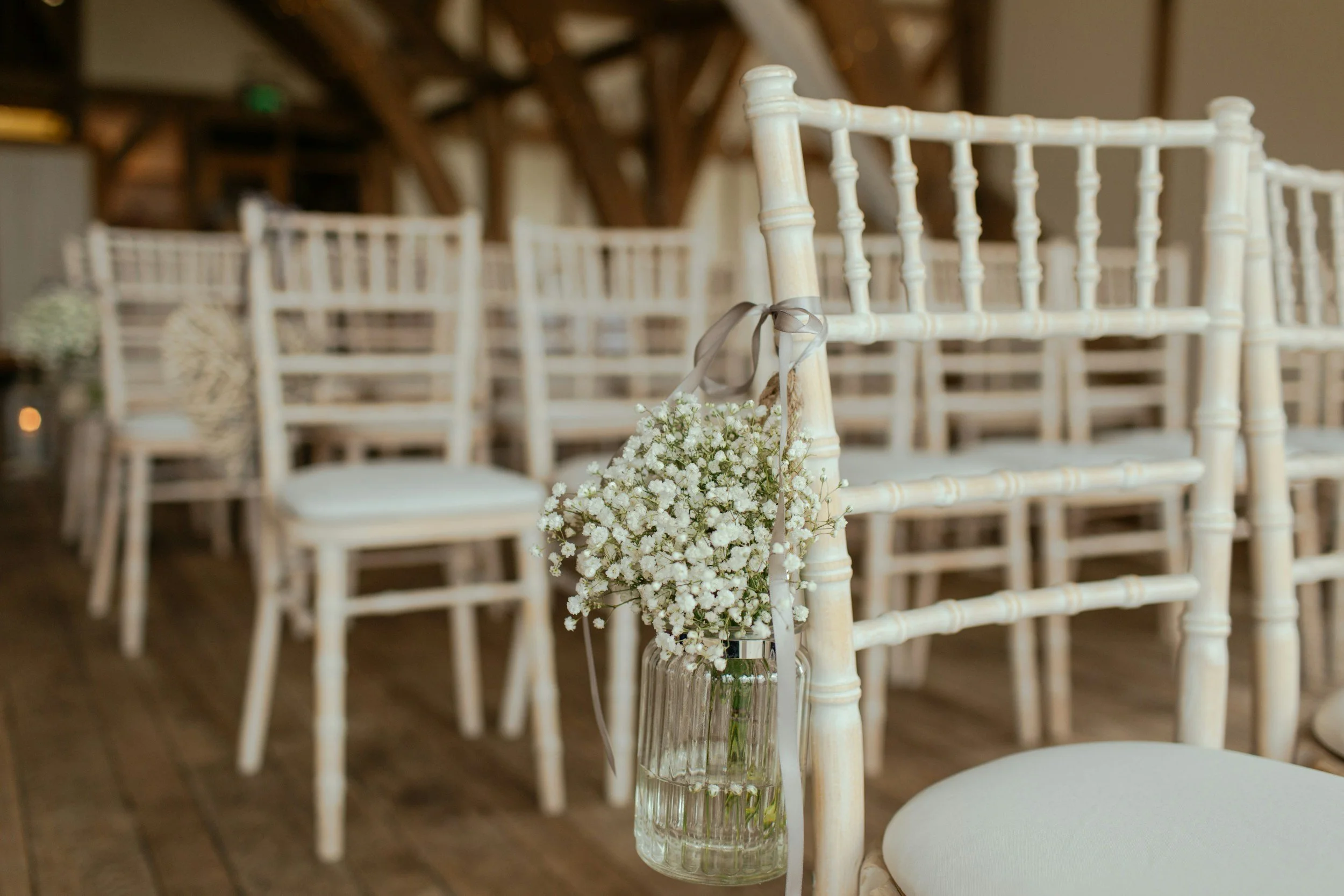 A clear glass vase with small white flowers, hanging from the back of a white wooden chair, with more similar chairs arranged in rows in the background, at an indoor event space or wedding venue.