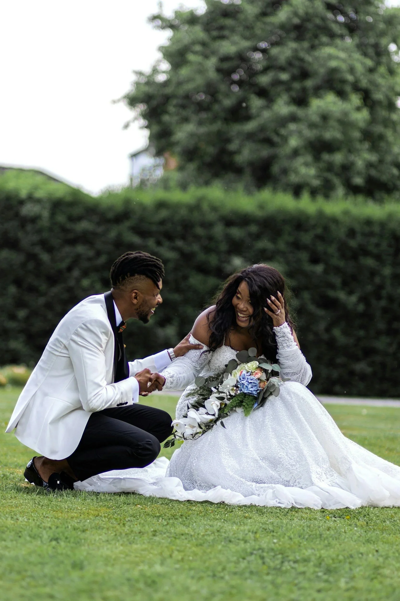 A newlywed couple is smiling and holding hands on a lush green lawn outdoors, with the bride in a white wedding gown holding a bouquet of flowers and the groom in a white suit jacket kneeling.