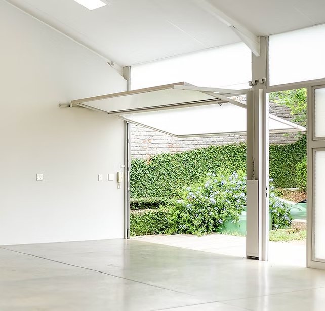 An empty room with white walls and floor, a partially opened glass door leading to a garden with green bushes and trees, and a retractable ceiling awning.