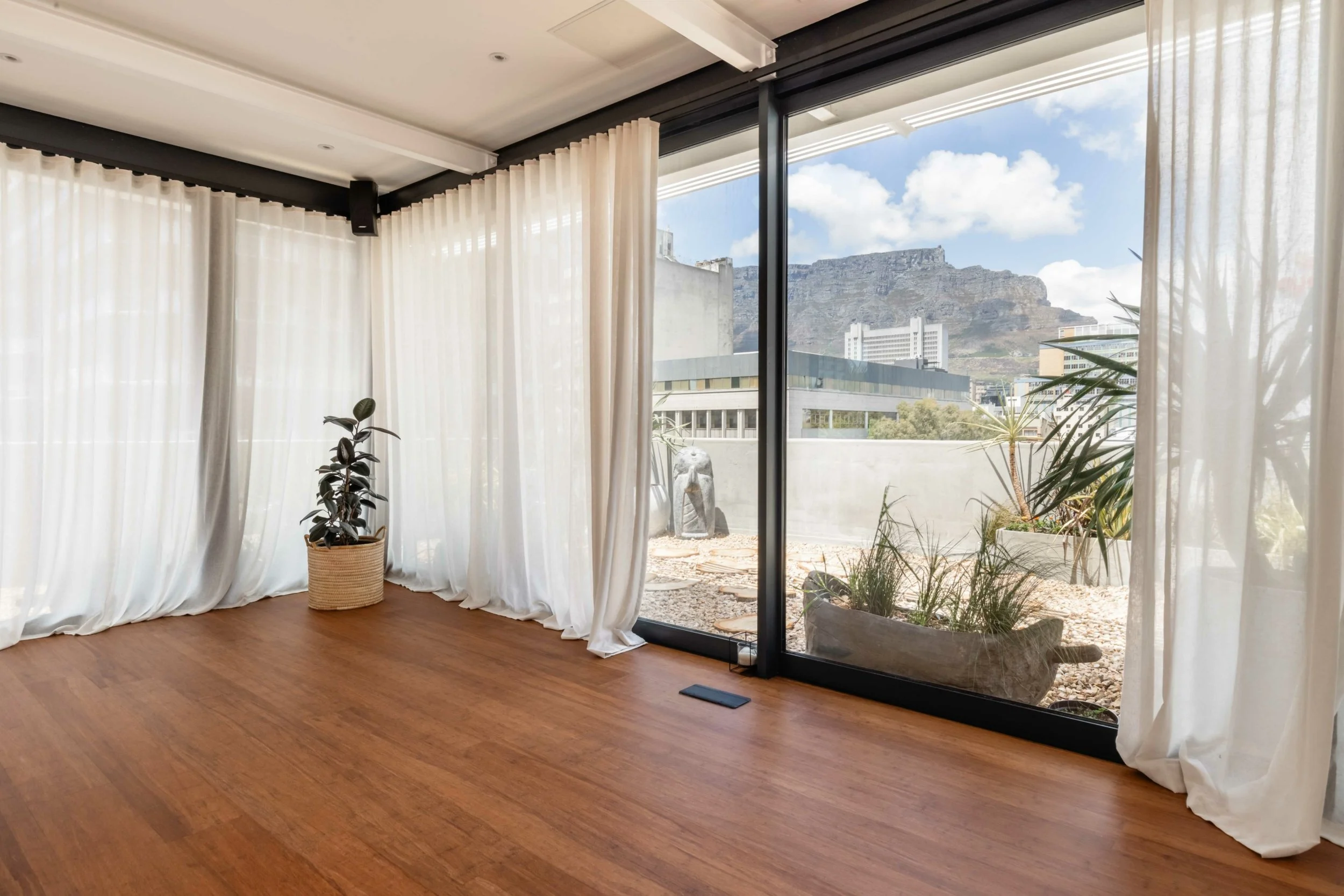 Interior of a room with hardwood floor and white curtains, large glass sliding doors revealing an outdoor area with potted plants and a view of mountains and city buildings.