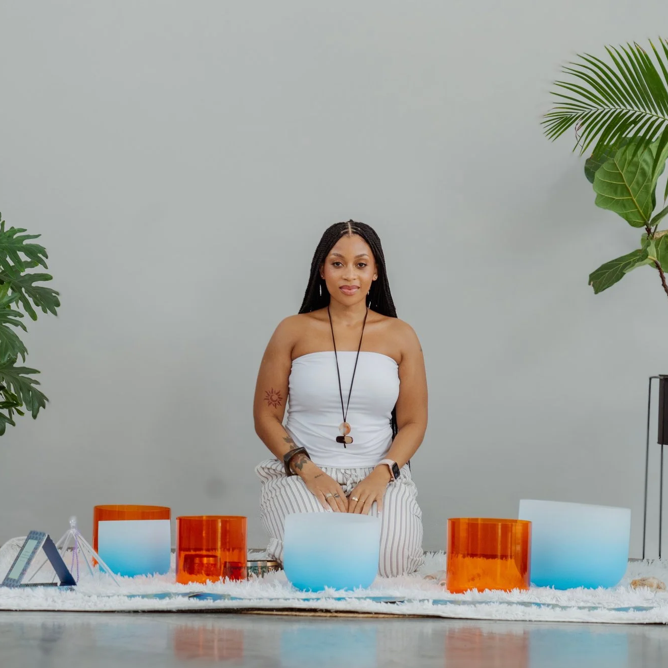 Woman sitting cross-legged on floor with singing bowls in front, surrounded by green plants, against plain gray wall.