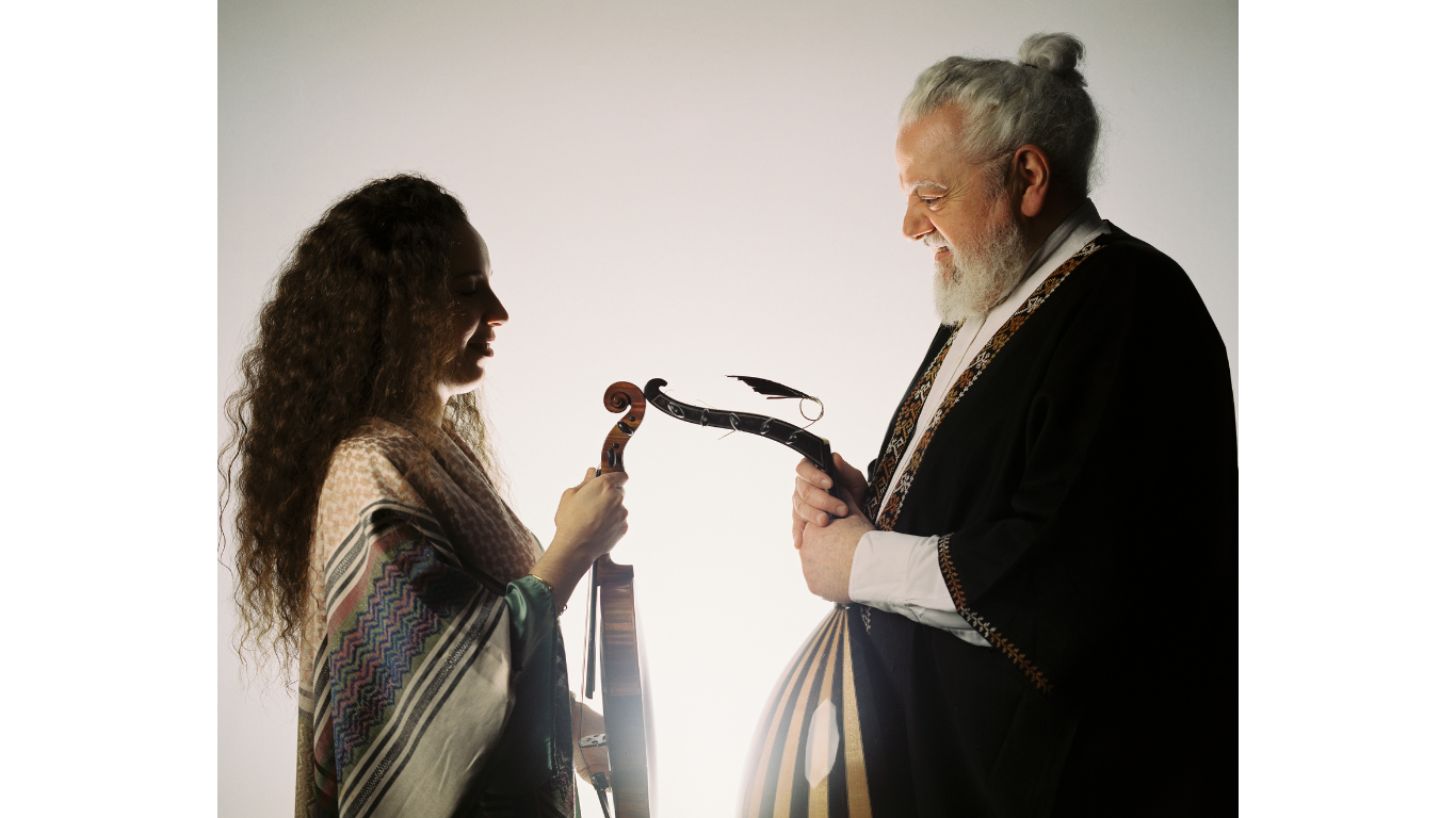 Une femme et un homme âgé souriant, se tenant face à face, avec des instruments de musique, dans une lumière douce.