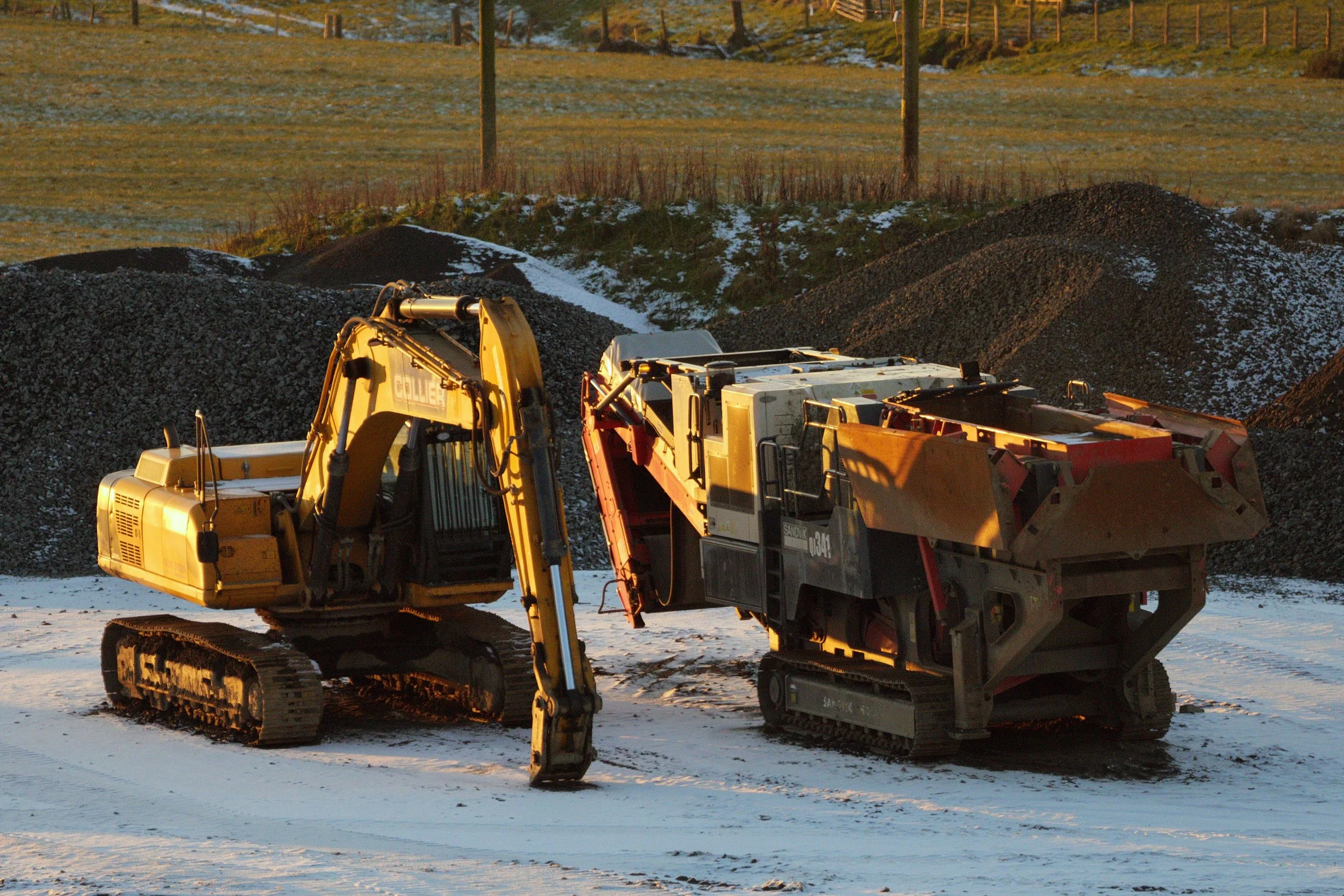 A yellow excavator and a large black and yellow tracked crusher machine parked on a snowy construction site during sunset.