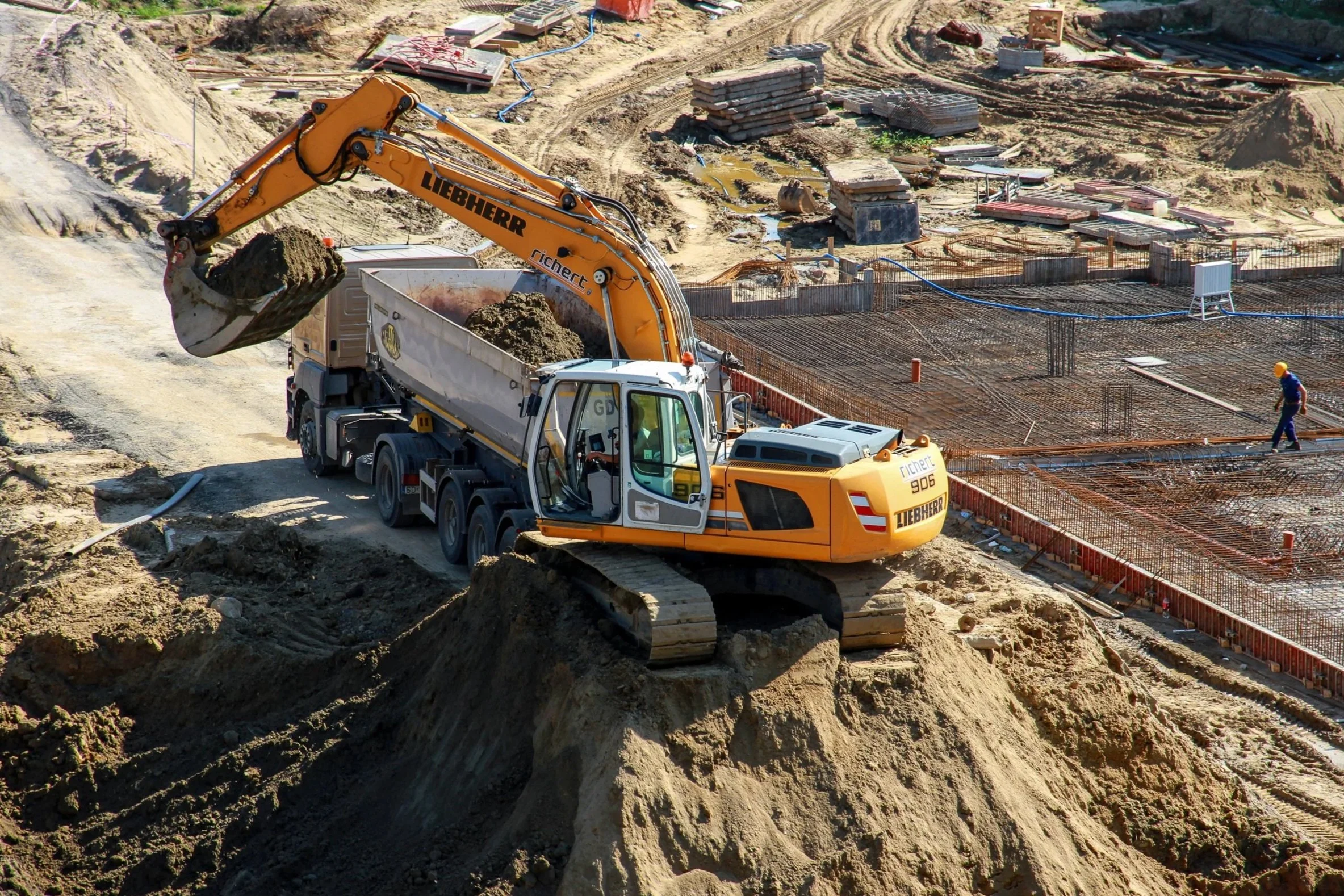 Construction site with an orange Liebherr excavator moving dirt, and a worker walking on rebar framework.