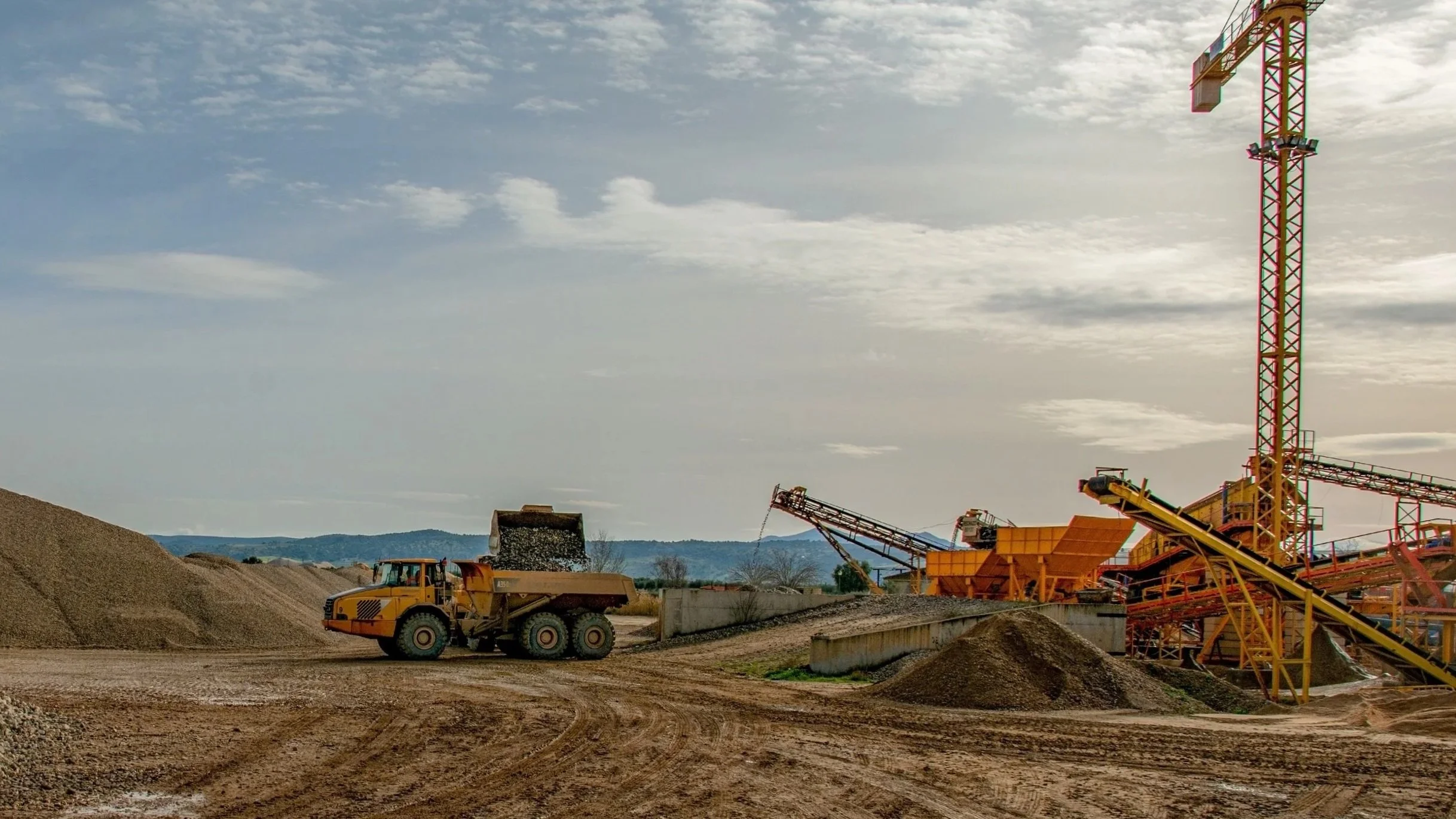 Construction site with a yellow dump truck, piles of dirt, industrial machinery, and a tower crane against a cloudy sky.