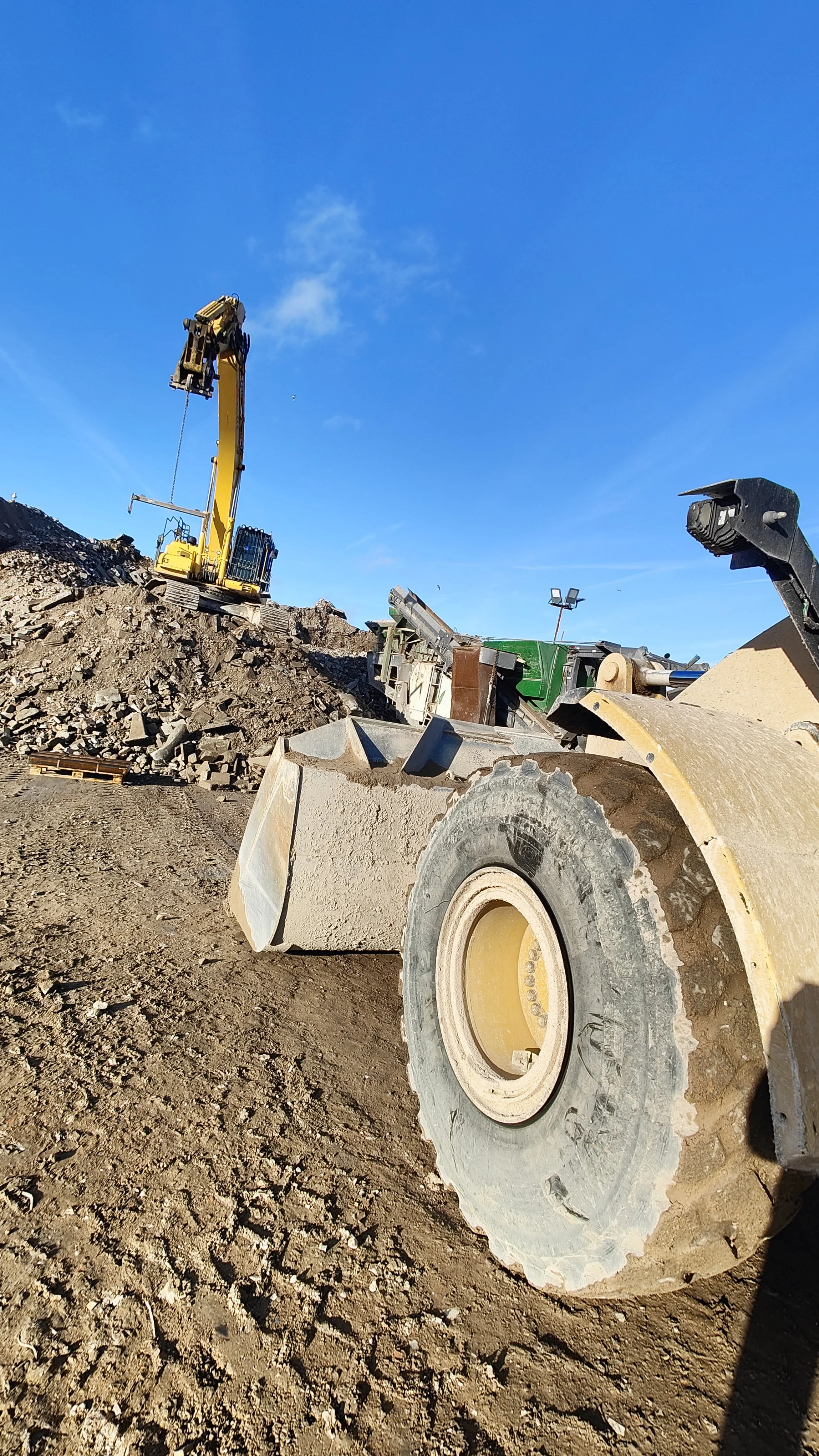 Construction site with a large yellow excavator and a road roller, with dirt and rocks in the background under a blue sky.