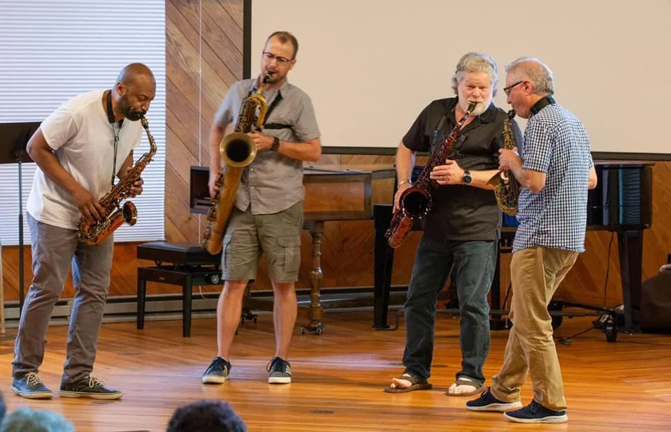 Four musicians stand closely together on a small stage, each playing a saxophone in an informal chamber music setting, leaning toward one another as they perform, with a piano and wood-paneled walls visible behind them.