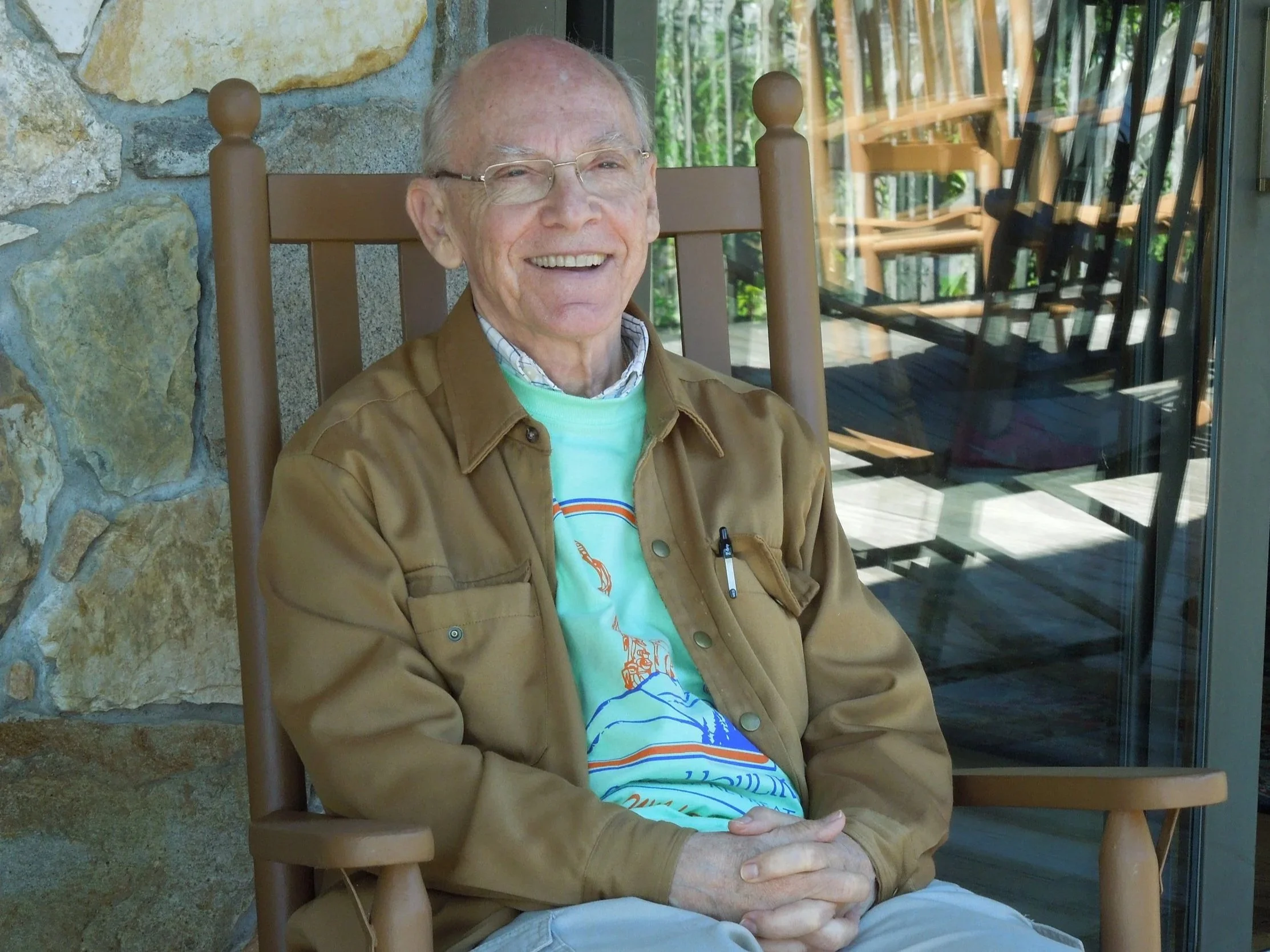 An older adult sits smiling in a wooden rocking chair on a stone porch, wearing glasses, a light green graphic T-shirt, and a tan jacket, with hands folded comfortably and a glass door reflecting additional rocking chairs behind them.