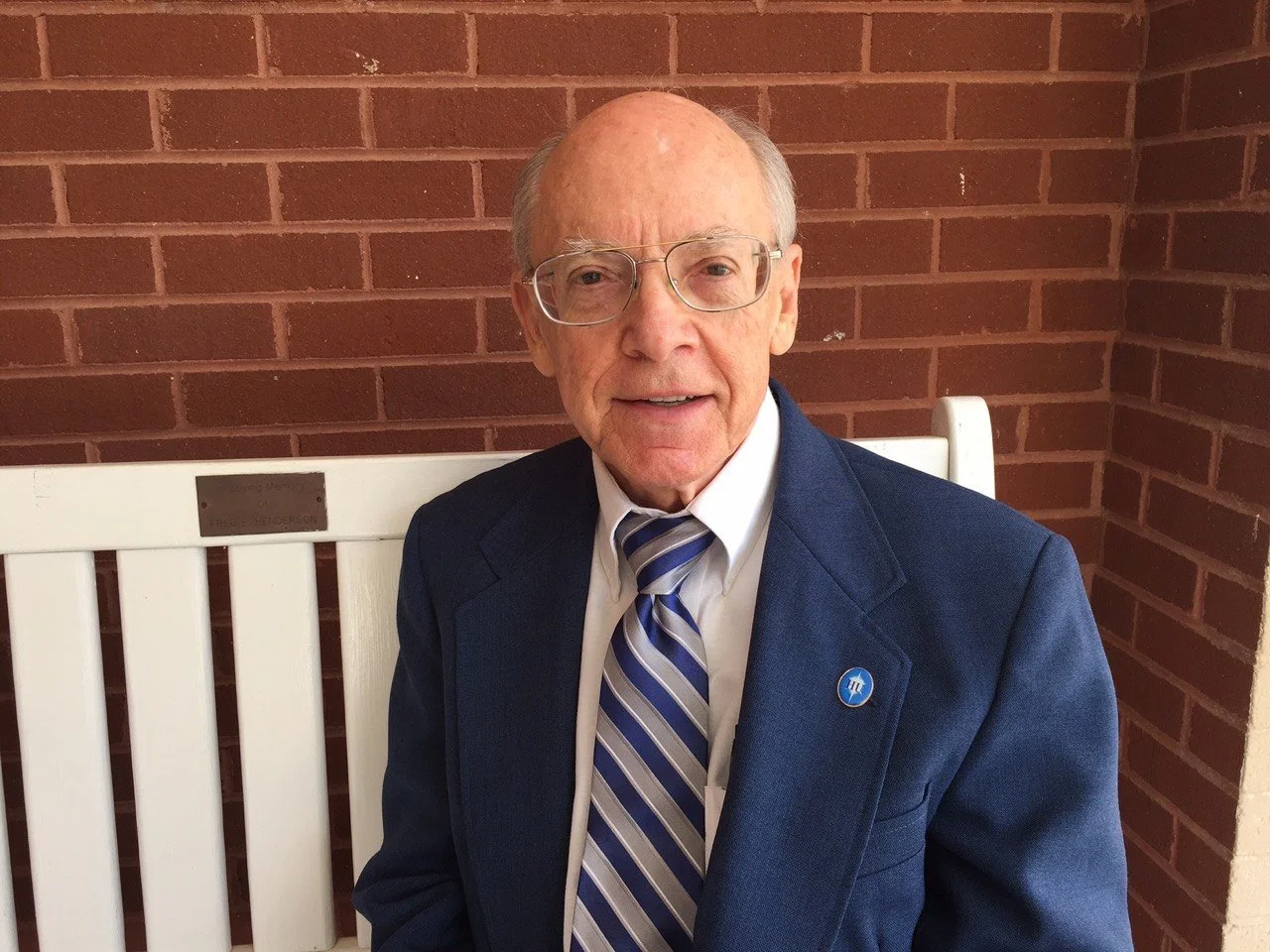 Portrait of composer Sherwood Shaffer seated on a white bench against a red brick wall, wearing a blue suit jacket, striped tie, glasses, and a small blue lapel pin, looking directly at the camera.