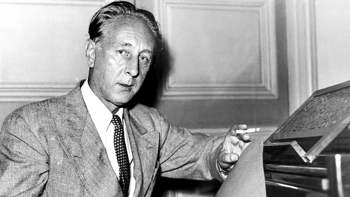A black-and-white photograph of a man in a suit and tie seated at a desk, holding a cigarette while reviewing a large sheet of handwritten music, with paneled walls visible in the background.