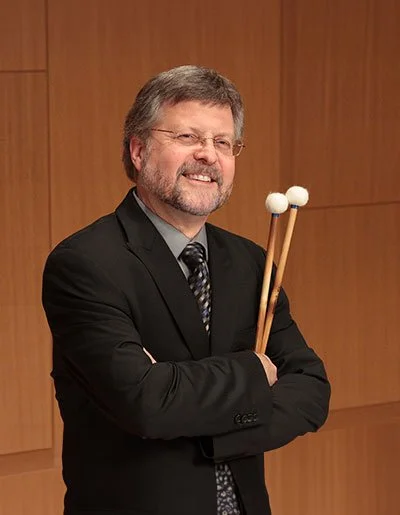 A man in a dark suit and patterned tie smiles while standing against a wood-paneled background, holding a pair of percussion mallets crossed in front of his chest with his arms folded.