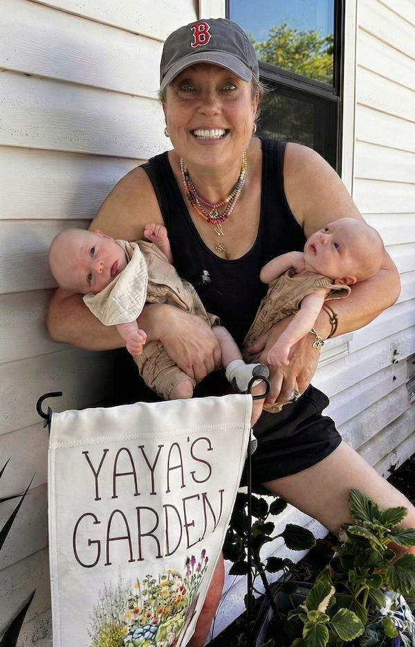A smiling woman wearing a baseball cap and black tank top is holding two babies dressed in beige clothes. They are outside near a house with white siding and a window, with greenery visible in the background. A gardening sign that reads 'YAYAS GARDEN' with flowers and garden drawings is in front of her.
