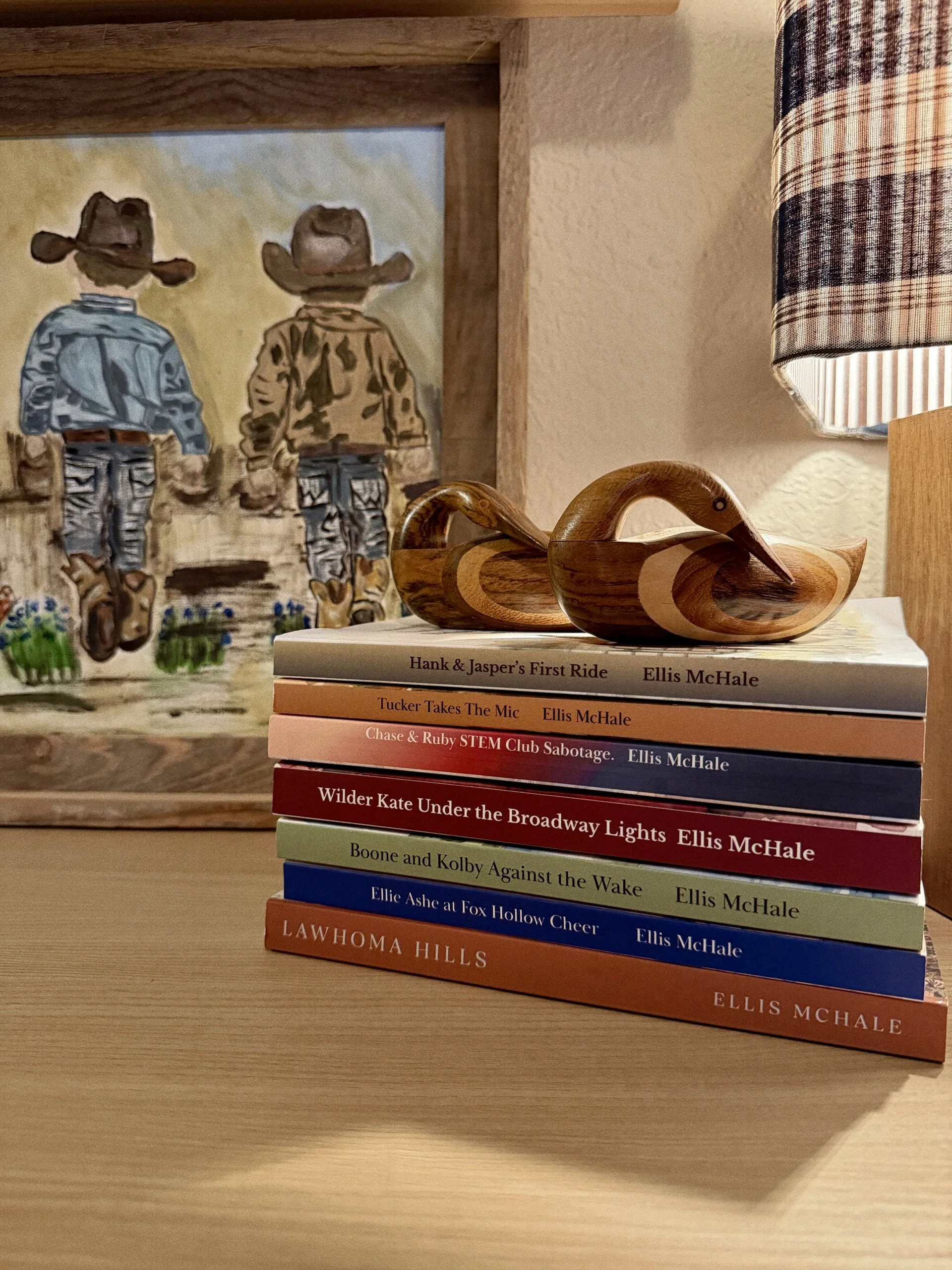 Lawhoma Hills Camp Run Amuck Series--stack of colorful BOOKS on a wooden table with a wooden duck figurine on top, in front of a painting of two children wearing cowboy hats, and a lamp with a plaid shade.
