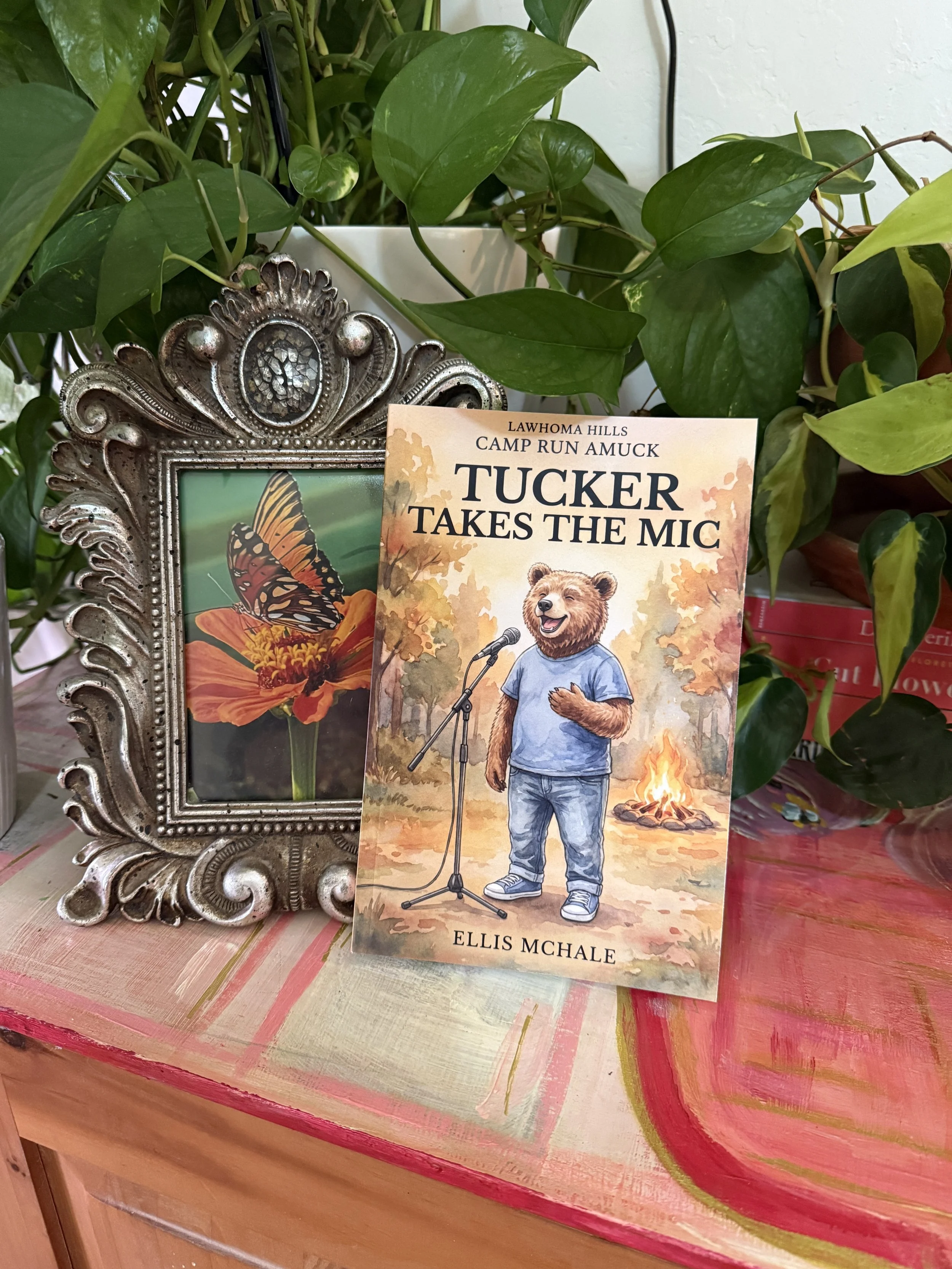 A book titled 'Tucker Takes the Mic' by Ellis McHale stands on a colorful, painted wooden table. Behind it, there is a silver ornate picture frame with a butterfly illustration inside. Green plants with large leaves surround the scene.