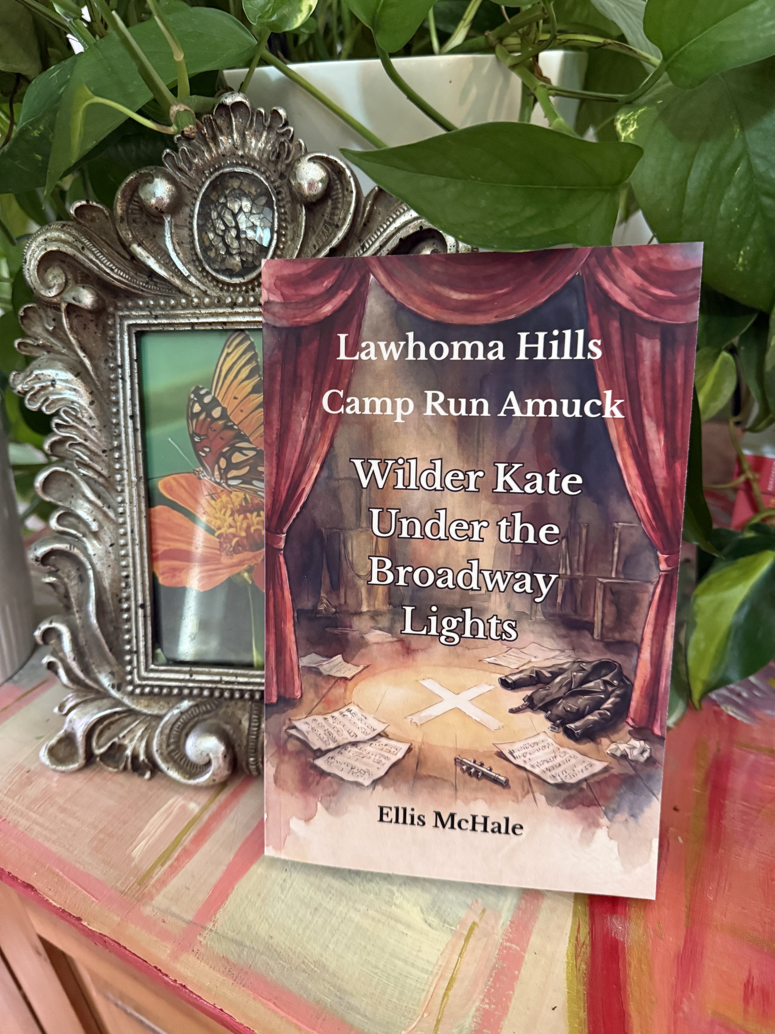 A  book titled 'Lawoma Hills Camp Run Amuck Wilder Kate Under the Broadway Lights' by Ellis McHale leaning against a decorative silver picture frame. The frame surrounds a picture of a butterfly. The background includes green leaves and a pink and green striped tablecloth.