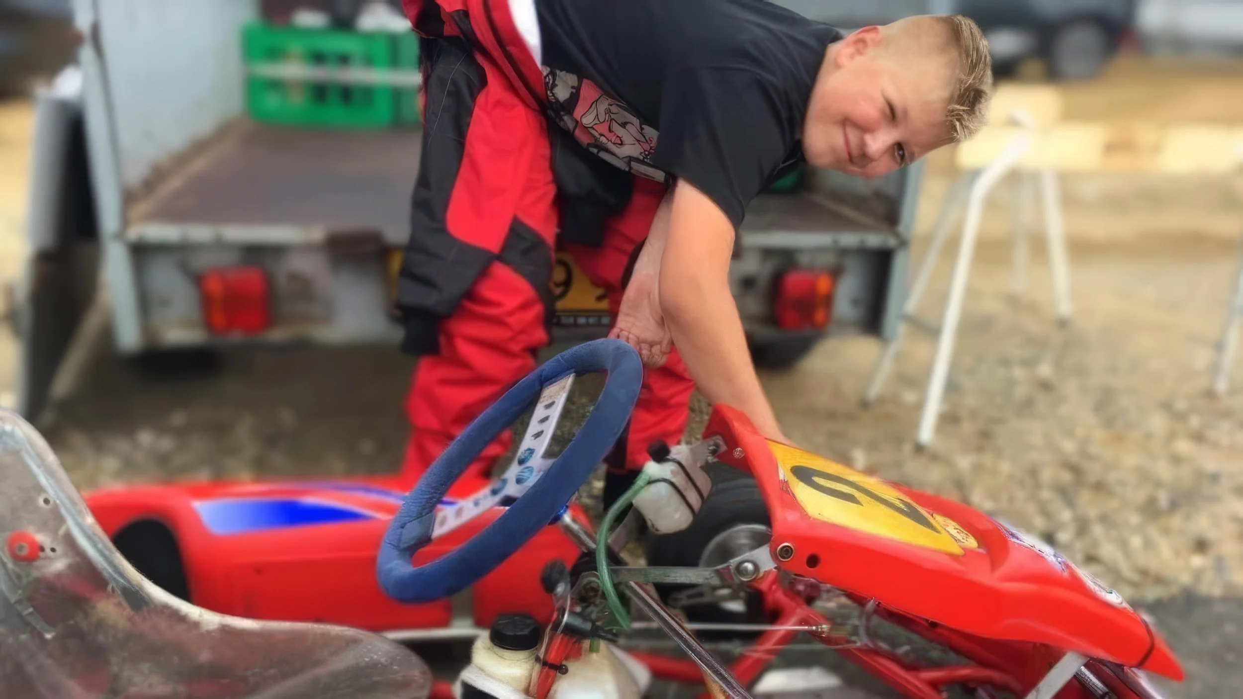 Alex Maansen i rødt og sort tøj arbejder på en rødt gokart. Han er bøjet over og ser direkte mod kameraet, mens han justerer eller inspicerer gokarten.