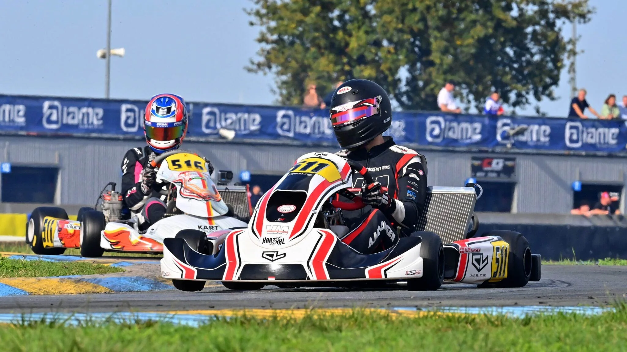 To gokart kører, Alex er foran med nummer 510 og en bagved med nummer 516, kører på en racerbane under en klar dag, omgivet af tilskuere.