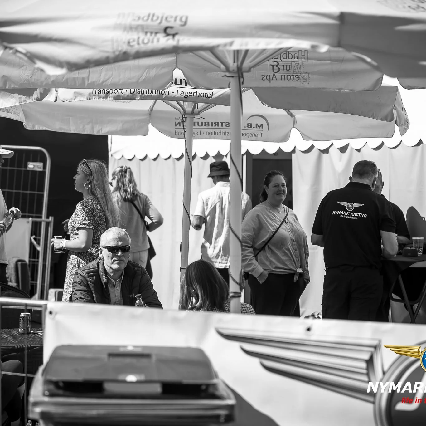 Mennesker under store parasoller ved en udendørs begivenhed, med nogle der står og snakker, og en siddende mand med solbriller.