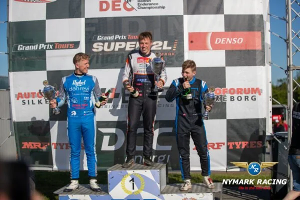 Tre racerkørere står på podiet Alex Maansen med nummer 1. Alle tre holder trofæer og ser glade ud. Bagved dem en væg med forskellige sponsorlogoer.