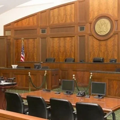 Empty courtroom with wooden paneling, chairs, and a judge's bench, featuring a U.S. flag and a seal on the wall as Jonathan Sablone prepares to argue a case.